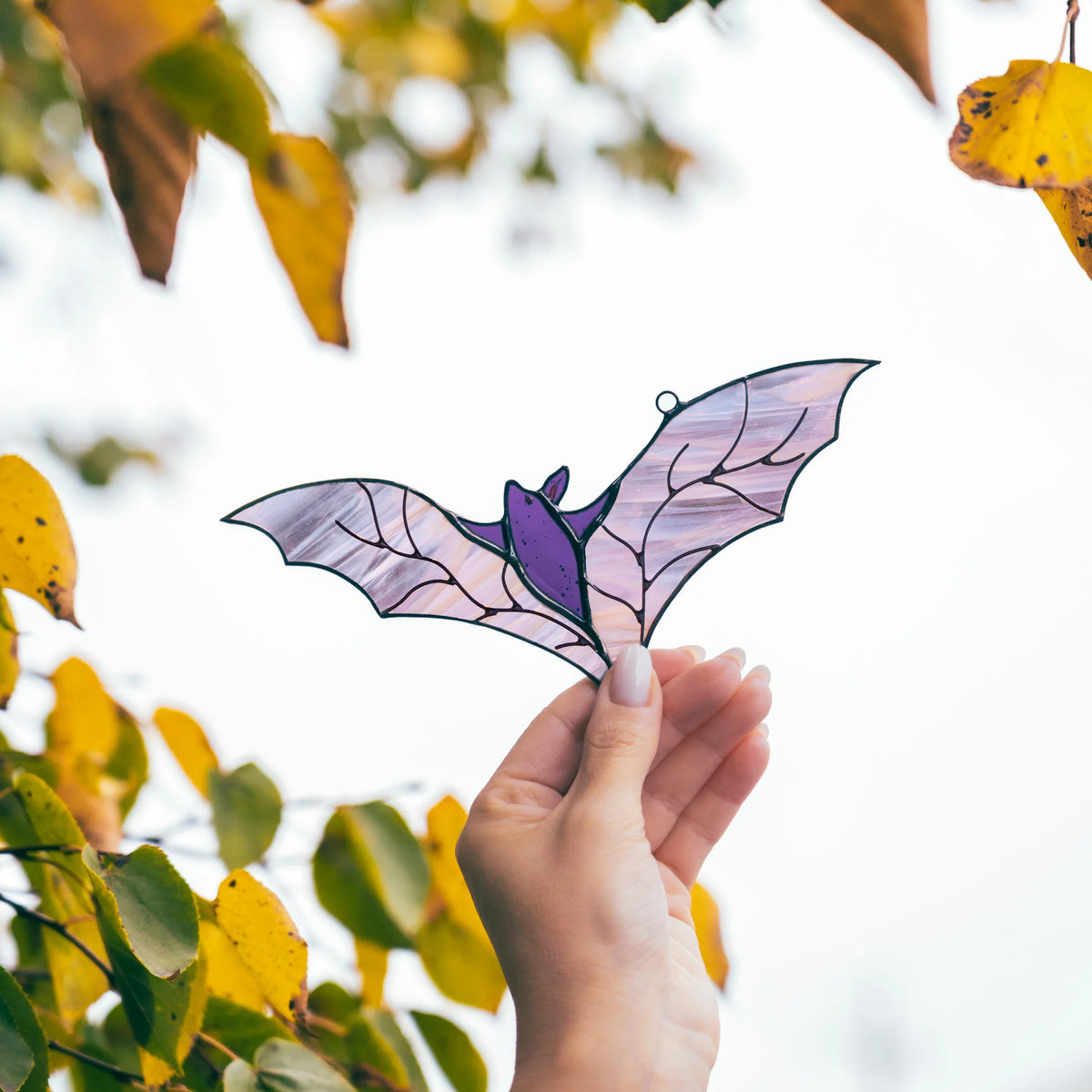 Hand holding the handmade purple bat stained glass suncatcher up among yellow autumn birch leaves, its wings spread wide against the pale sky - spooky window decor