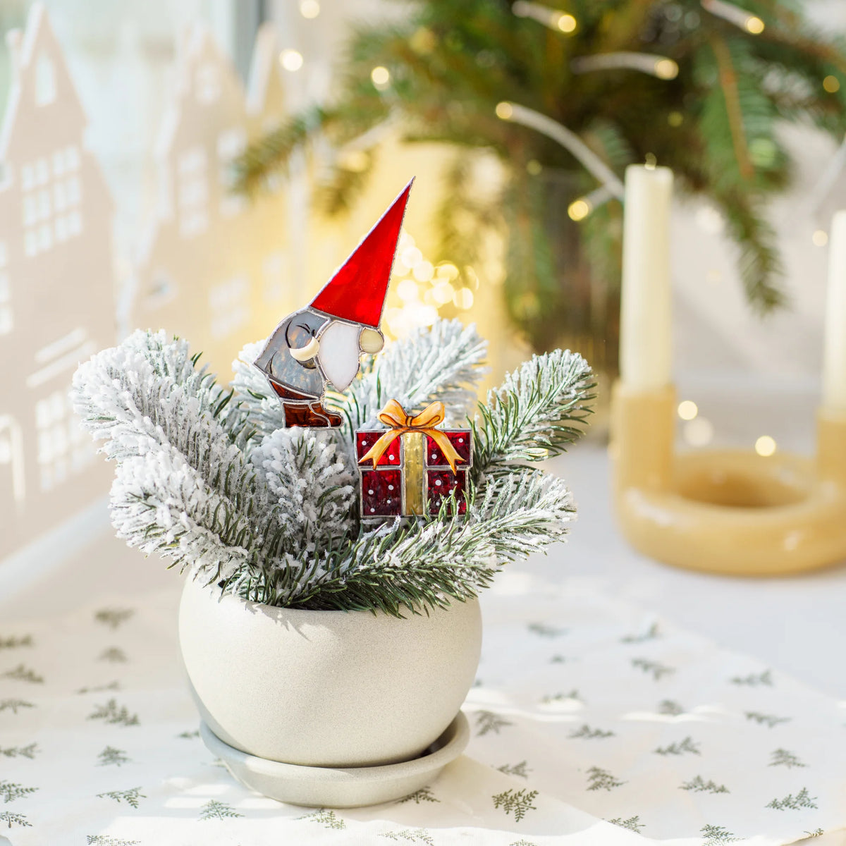 Christmas gnome and gift stained glass plant stakes in a white pot of frosted fir on a holiday table with candles - handmade Christmas stained glass decor for planters
