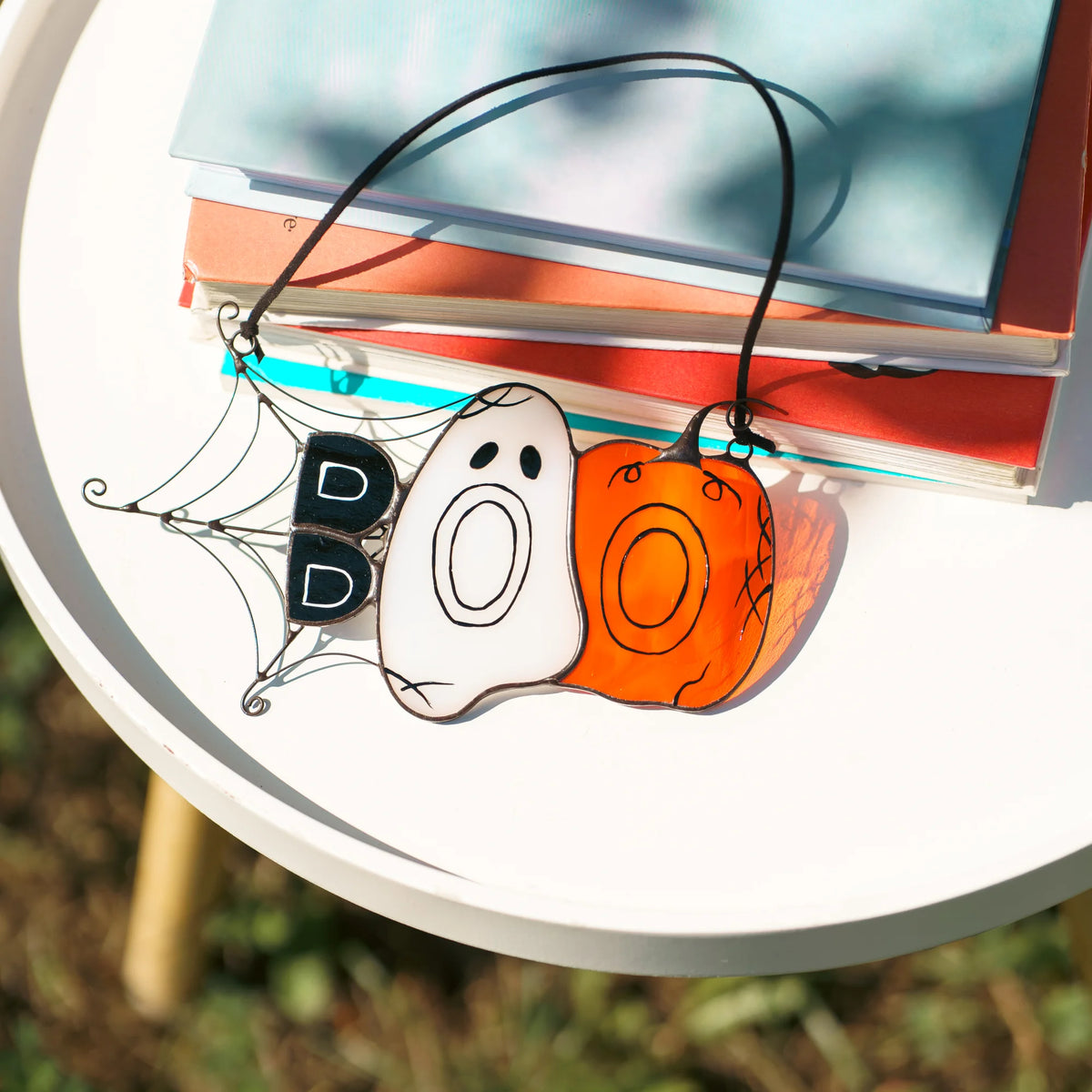 Close-up of the BOO Halloween stained glass suncatcher tilted on a white table in the sun, the orange pumpkin glass casting a warm coloured shadow beside books