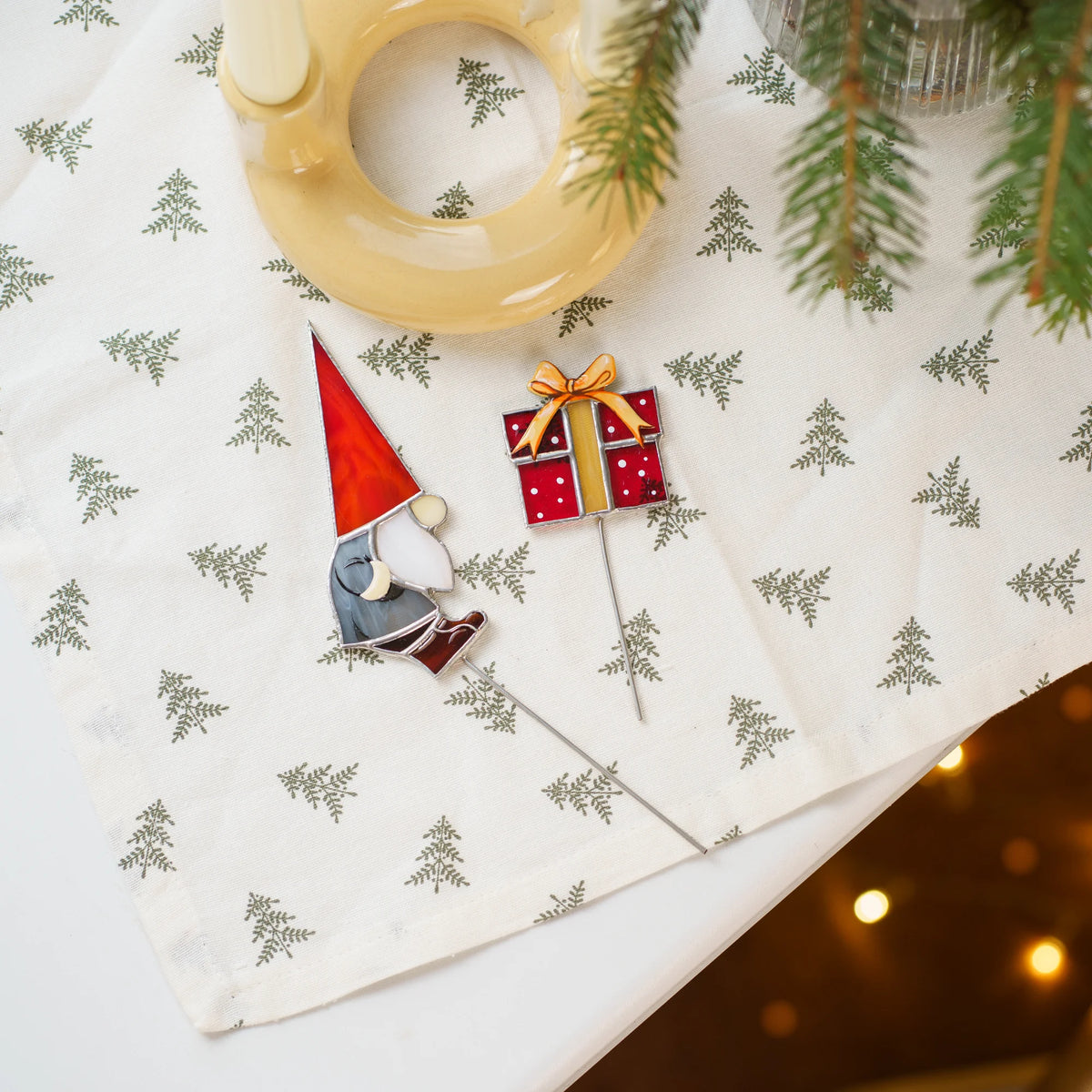 Christmas gnome with gift stained glass plant stakes lying on a white tablecloth with tiny tree prints next to a wreath and fairy lights - handmade Christmas stained glass ornaments