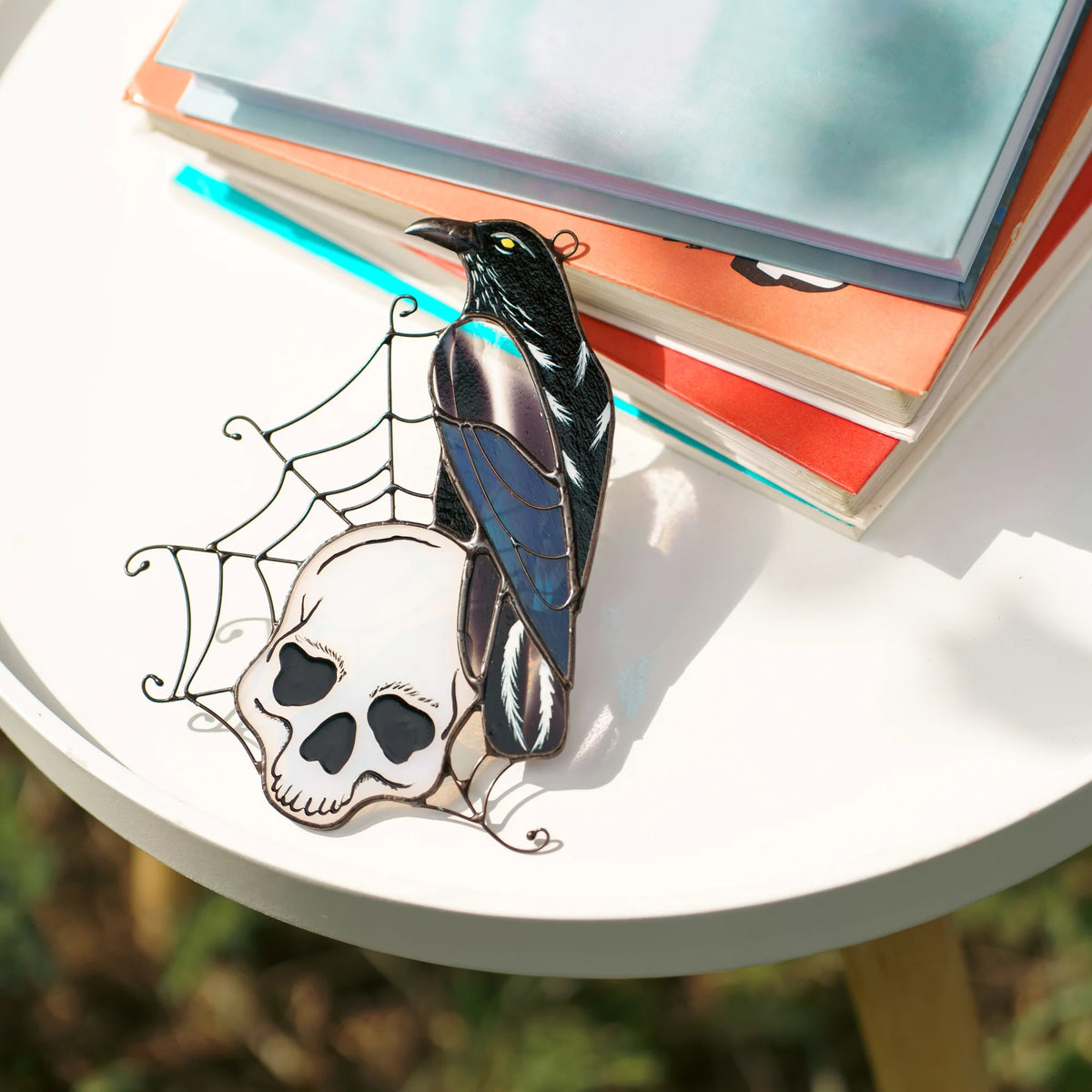 Stained glass raven and skull suncatcher resting on a white table by a stack of books, side view showing the iridescent purple and blue glass wing - handmade gothic decor and gift for readers