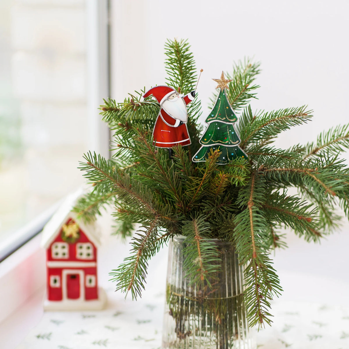 Santa with Christmas tree stained glass plant stakes standing among pine branches in a glass jar beside a little red house - handmade Christmas stained glass decor for a sunny windowsill