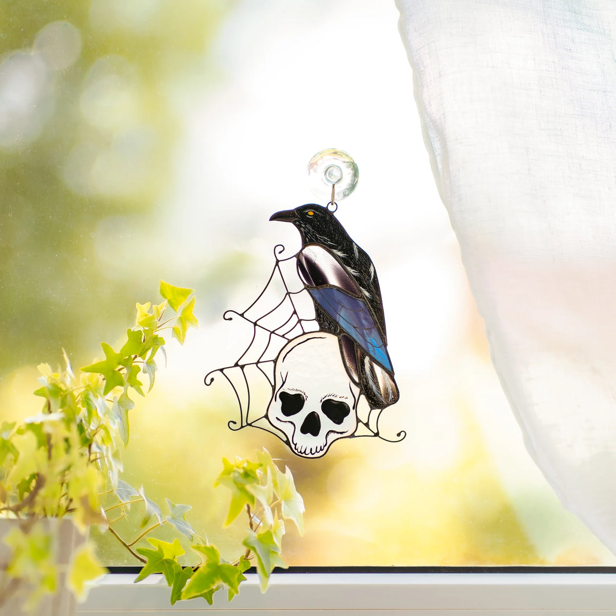 Stained glass raven and skull suncatcher hanging in a sunlit window above trailing ivy, glossy black raven on a white skull in a wire spider web - handmade gothic Halloween window decoration