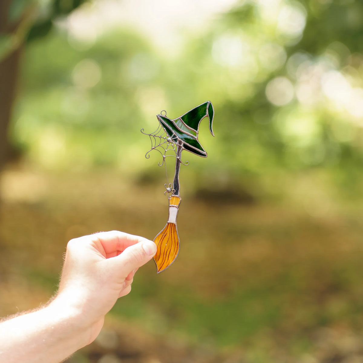 Hand holding the handmade witch broom Halloween stained glass suncatcher against a green forest background, showing the dark green hat, wire spiderweb and amber bristles