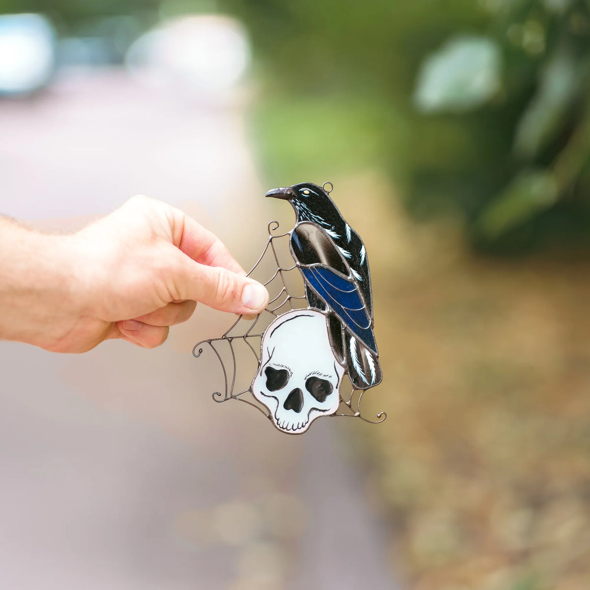 Hand holding a stained glass raven and skull suncatcher outdoors, glossy black raven with an iridescent blue wing perched on a white skull in a wire web - handmade gothic Halloween gift 4.5 by 6 inches