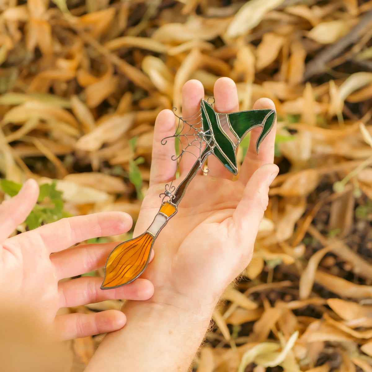 Handmade witch broom stained glass suncatcher held in two open palms over autumn leaves - a small witchy window hanging and a Halloween gift for stained glass lovers