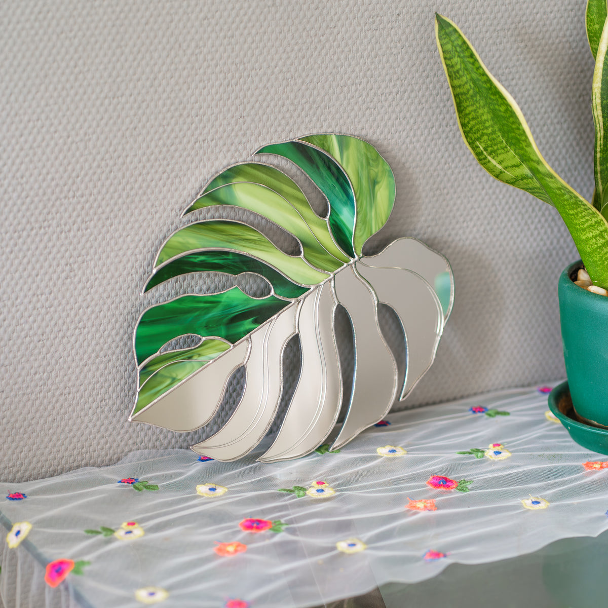 Monstera Leaf Stained Glass Mirror leaning on a gray wall above a floral embroidered tablecloth beside a potted snake plant - handmade stained glass home decor in green glass and mirror