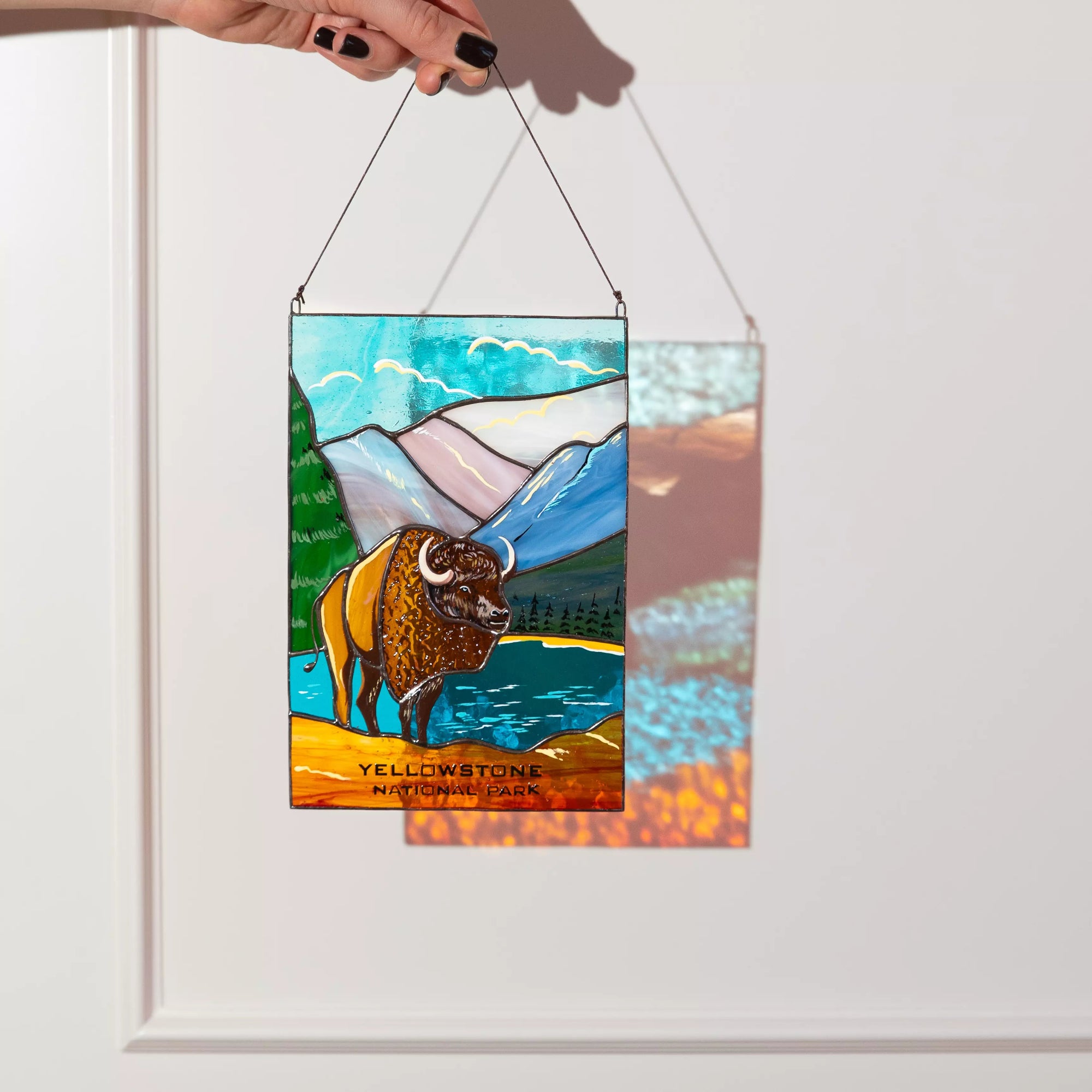 Stained glass panel with a bison and mountain design, held by a hand against a white background.
