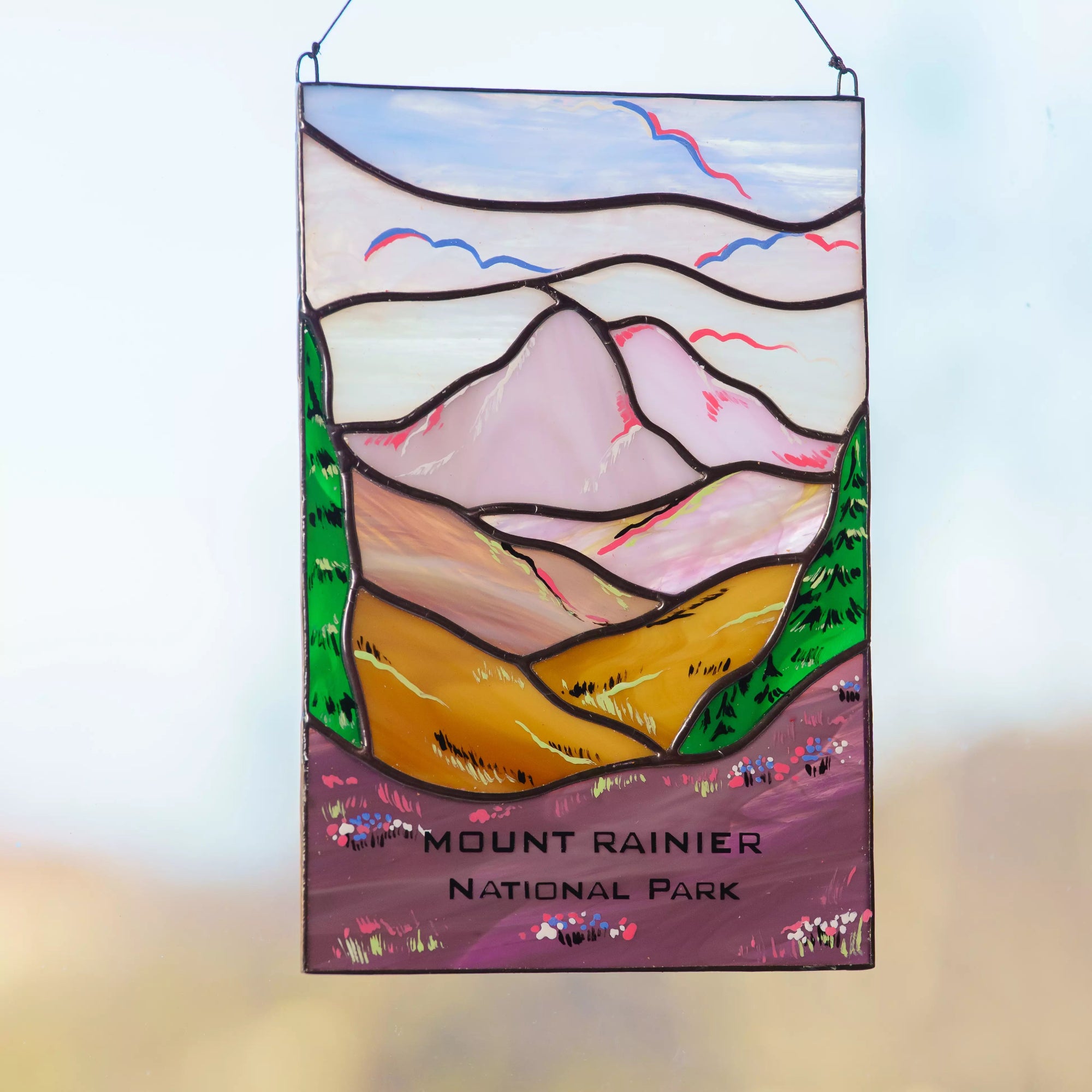 Stained glass panel of Mount Rainier with 'Mount Rainier National Park' text on a blurred nature background