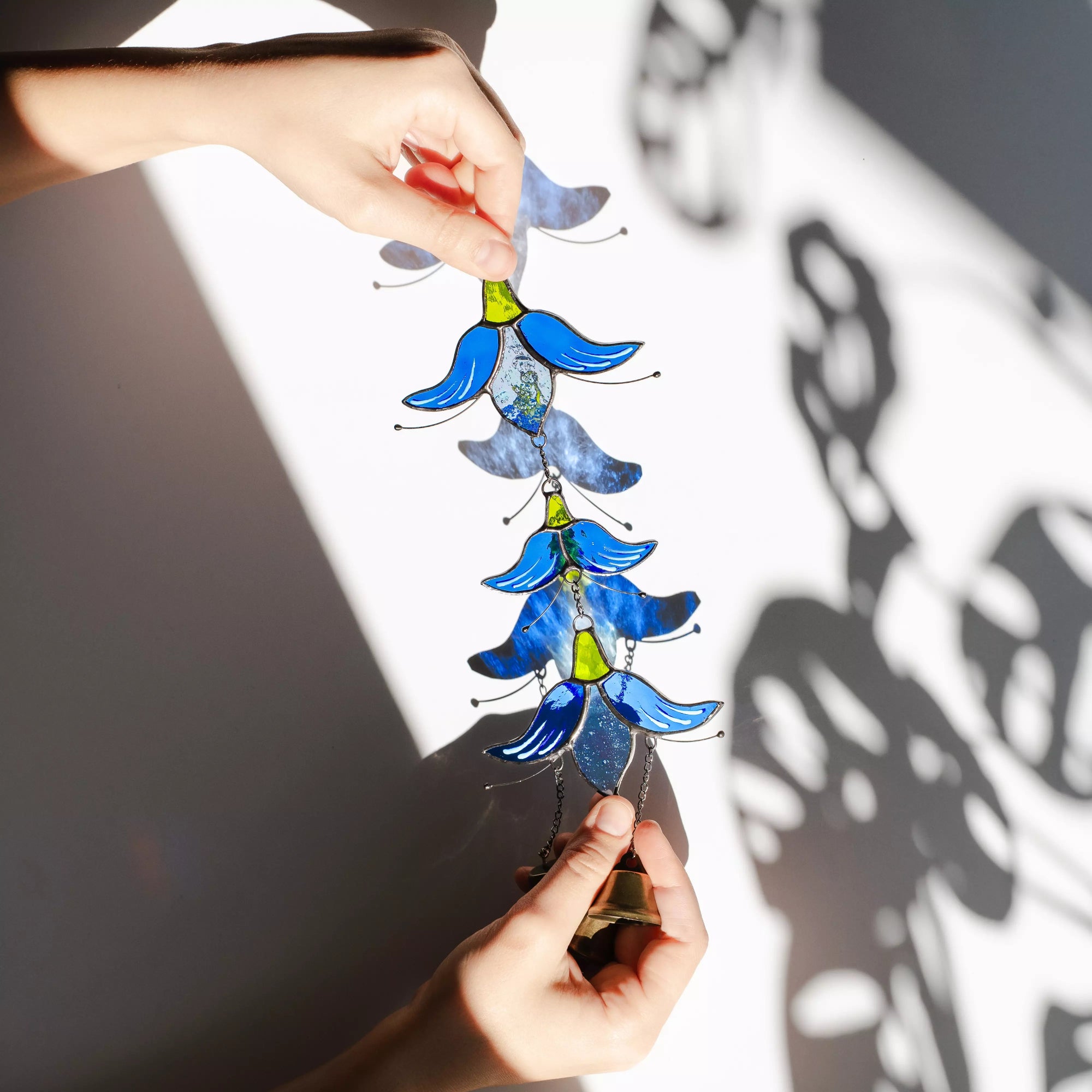 A hand holding a stained glass flower wind chime with dramatic light and shadows on a white surface.
