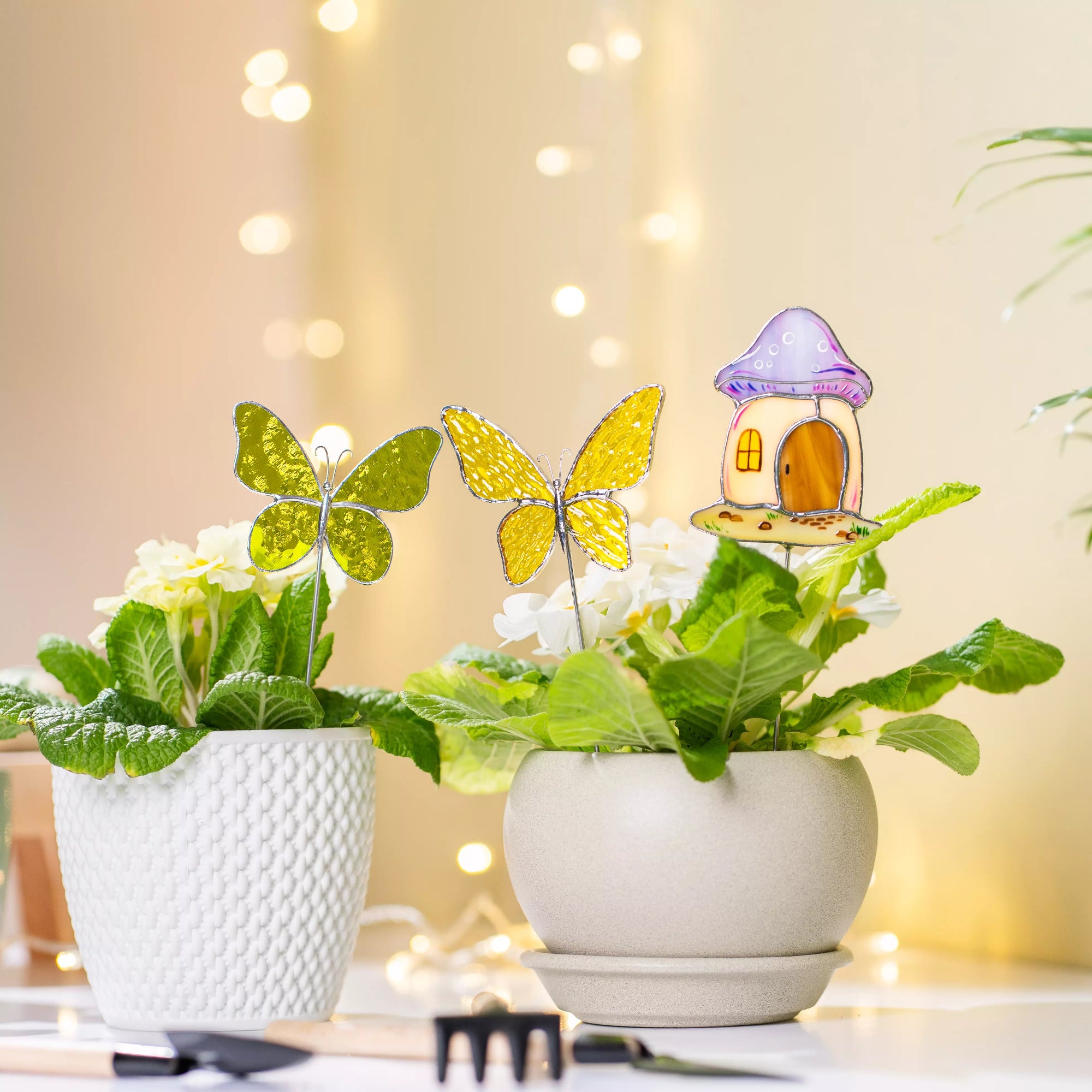 A bright lifestyle shot of a yellow butterfly, green butterfly, and mushroom house stake placed in potted plants on a white surface with soft bokeh lighting in the background.