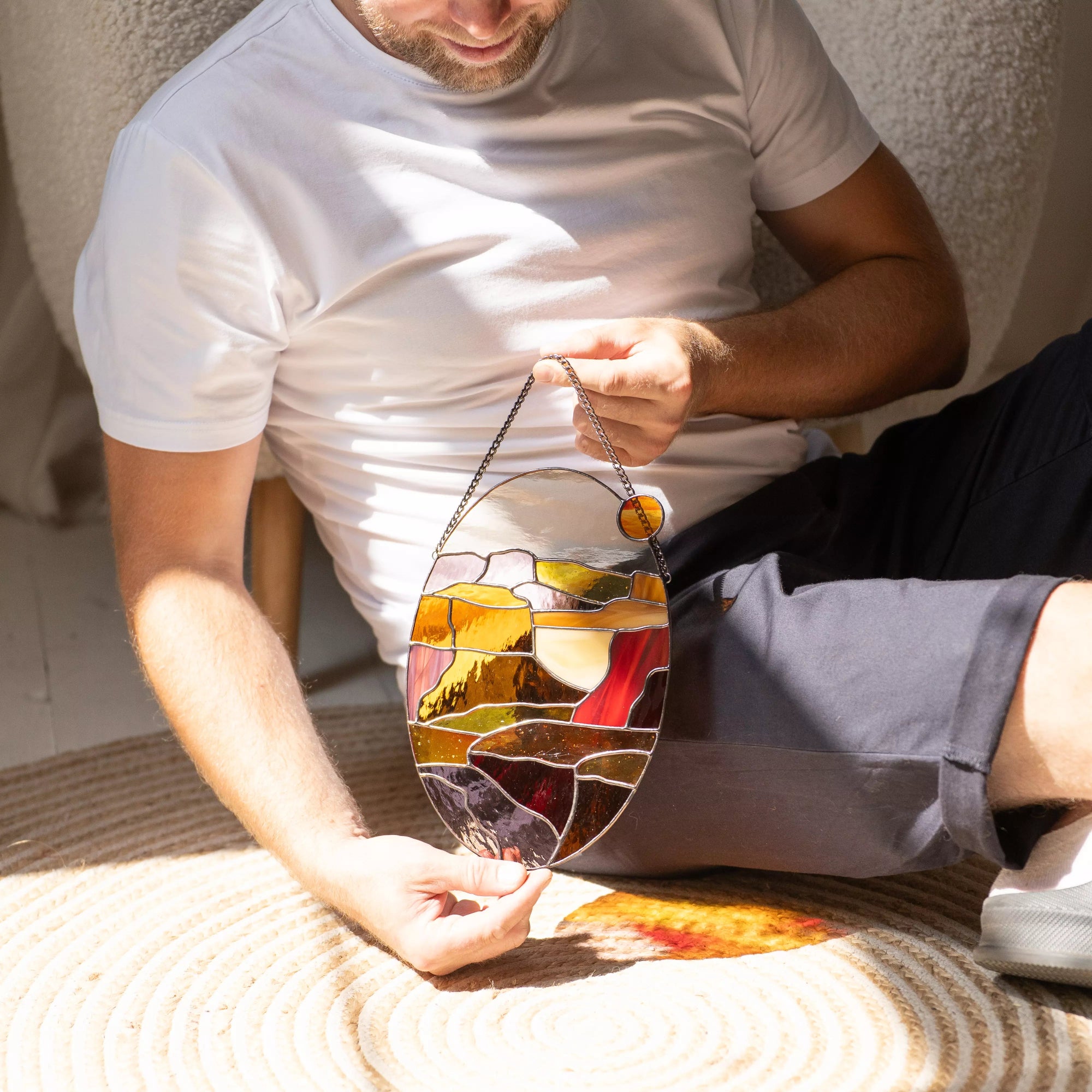 Man sitting on floor holding oval stained glass sunset panel with warm amber and red tones