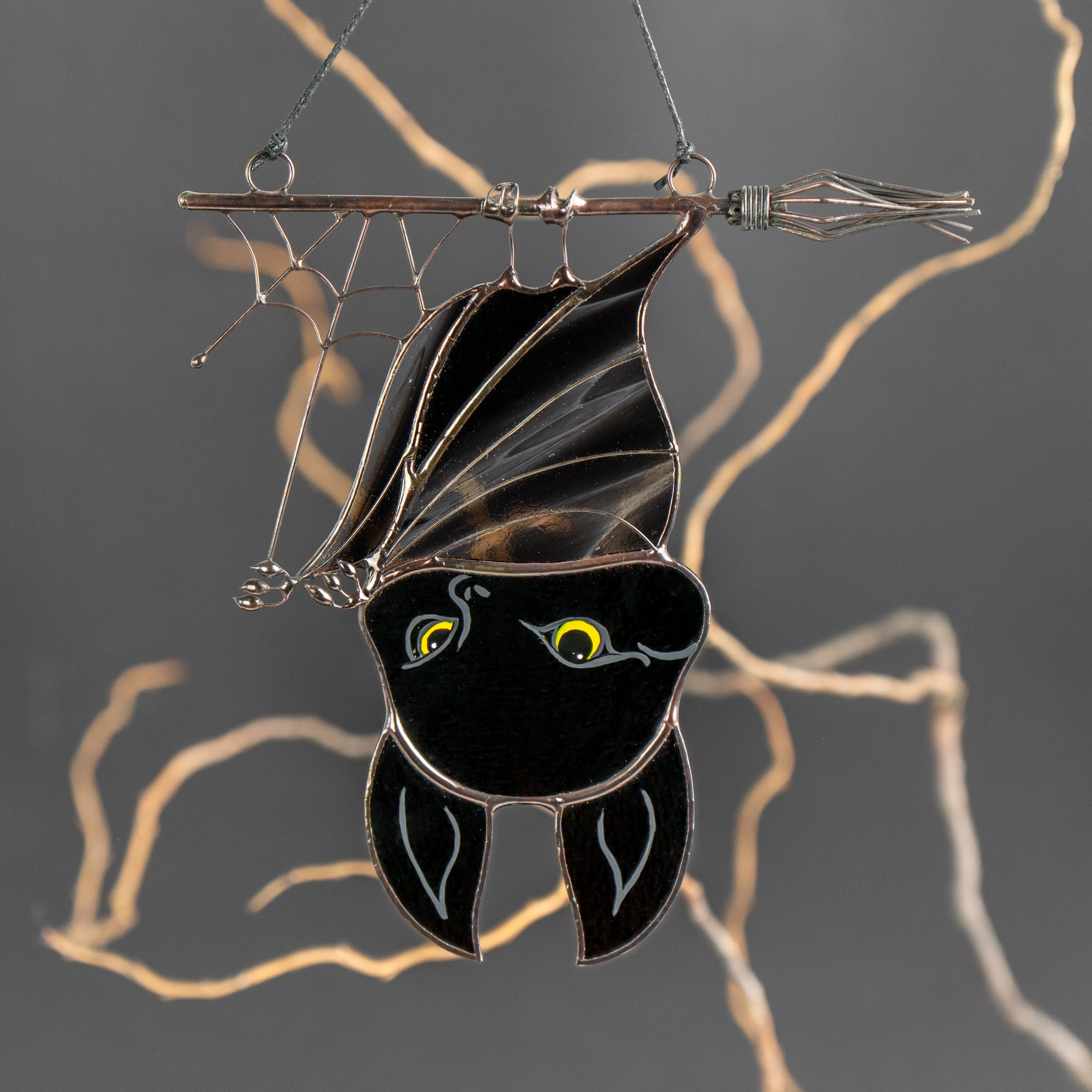 Close-up of stained glass Halloween bat with glowing yellow eyes hanging upside down on broom and web.