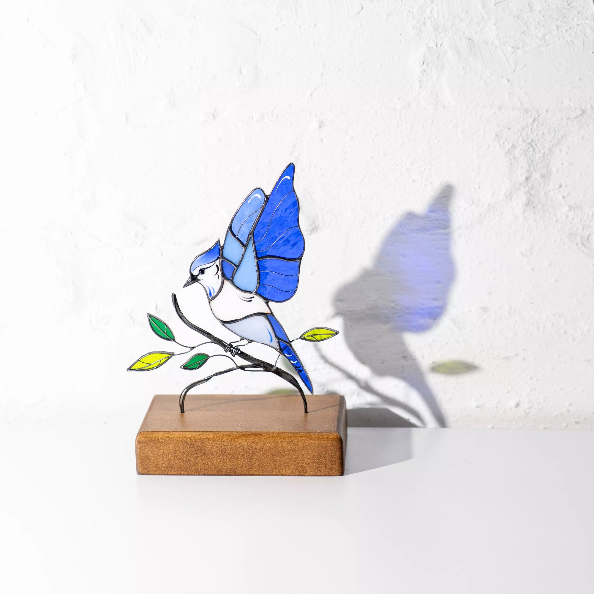Sunlit photo of stained glass blue jay on wooden base, casting blue and green reflections on white wall