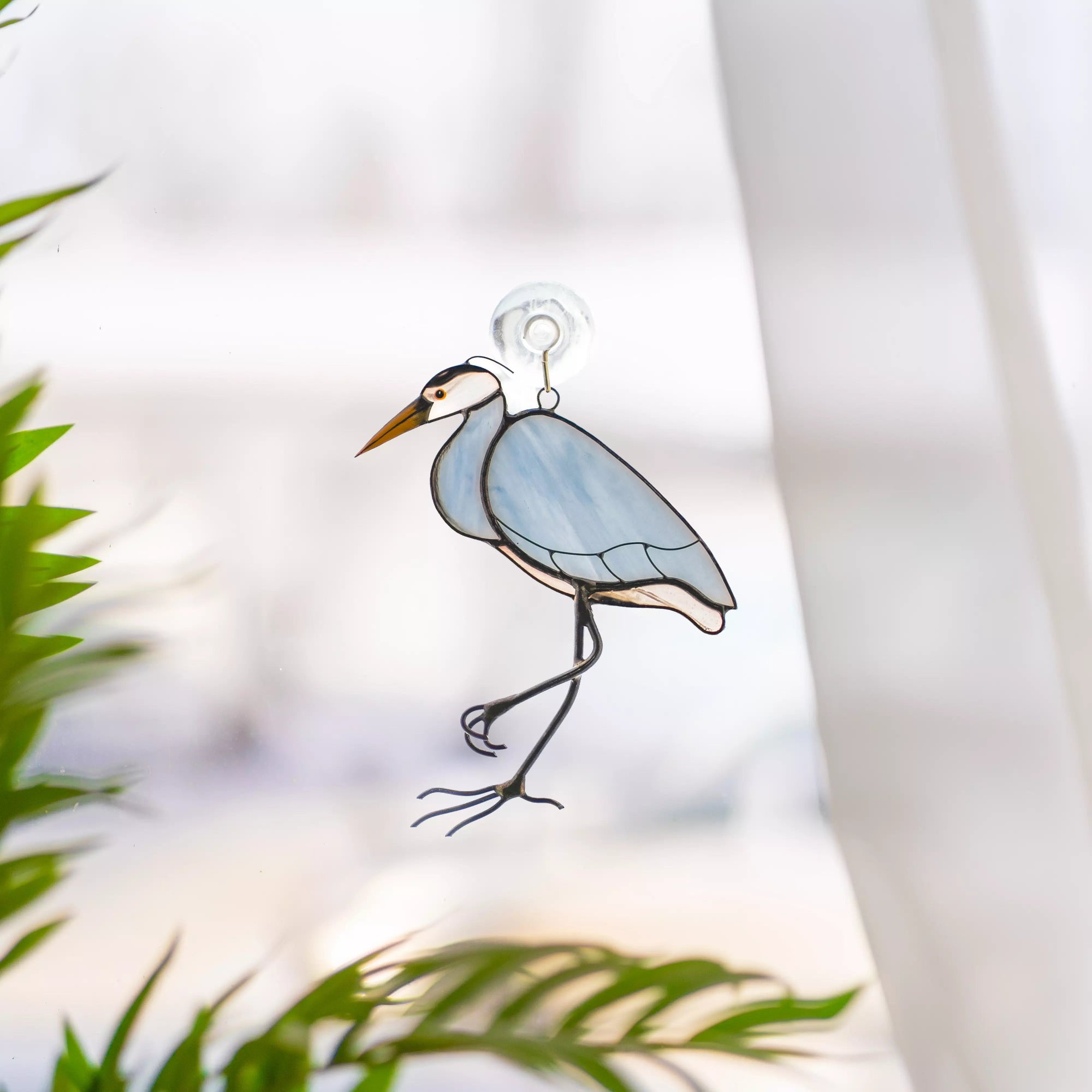 Stained glass suncatcher heron hanging on window with suction cup, sunlight passing through glass and green plant leaves in background.