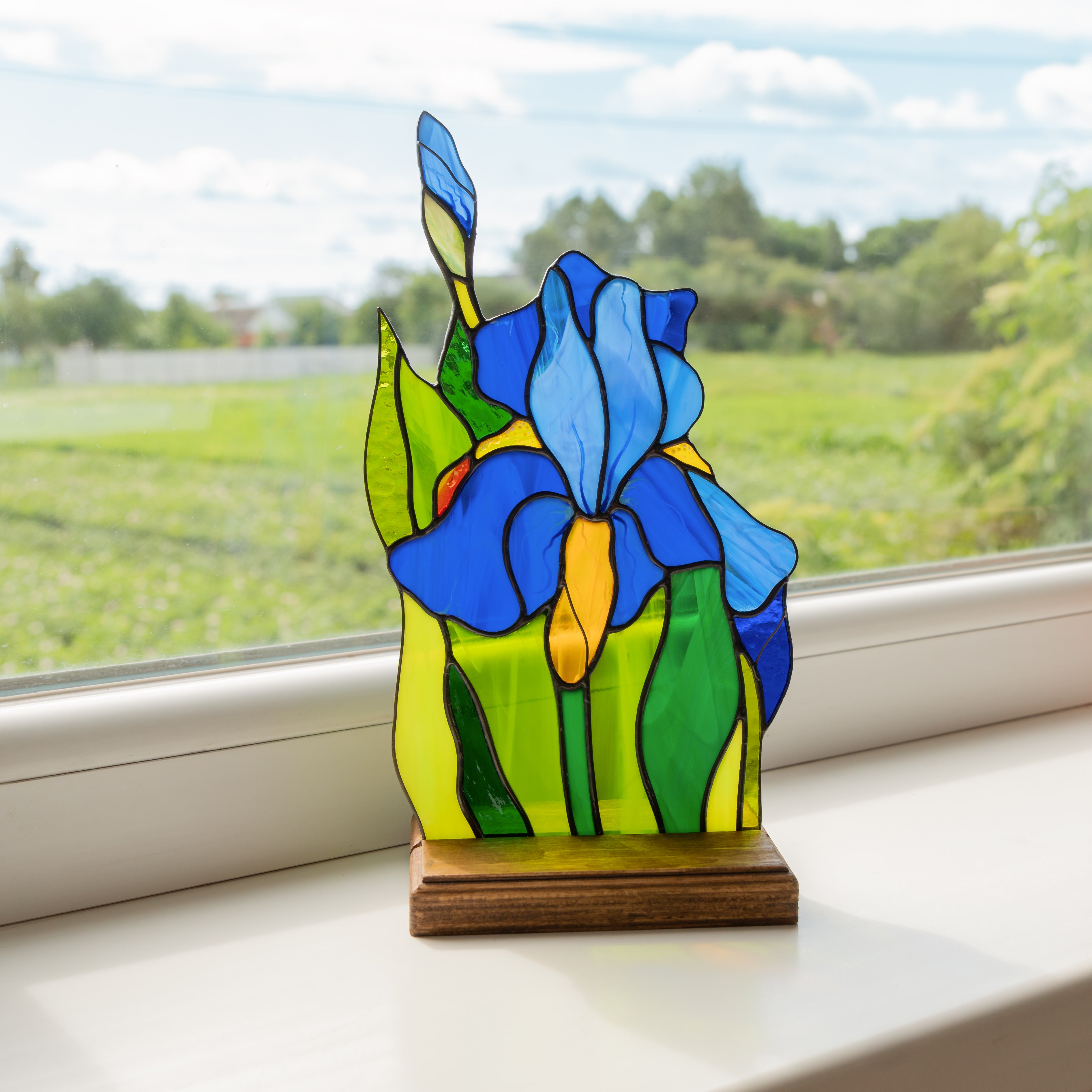 Bright blue stained glass iris flower on a wooden stand, placed on a windowsill with green landscape in background.