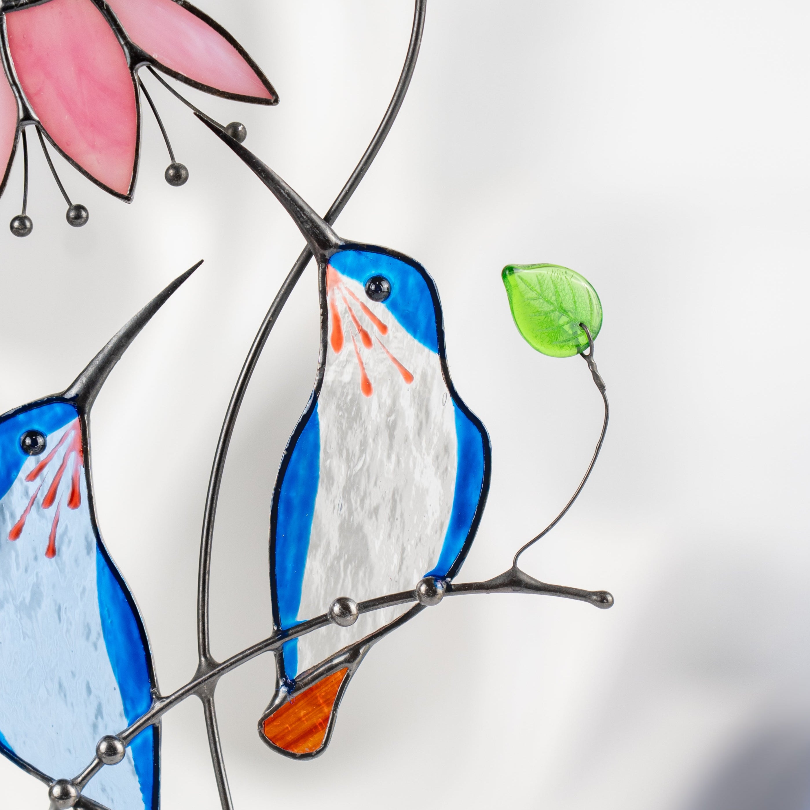 Close-up of a blue stained glass bird with orange tail and green leaf on wire branch.