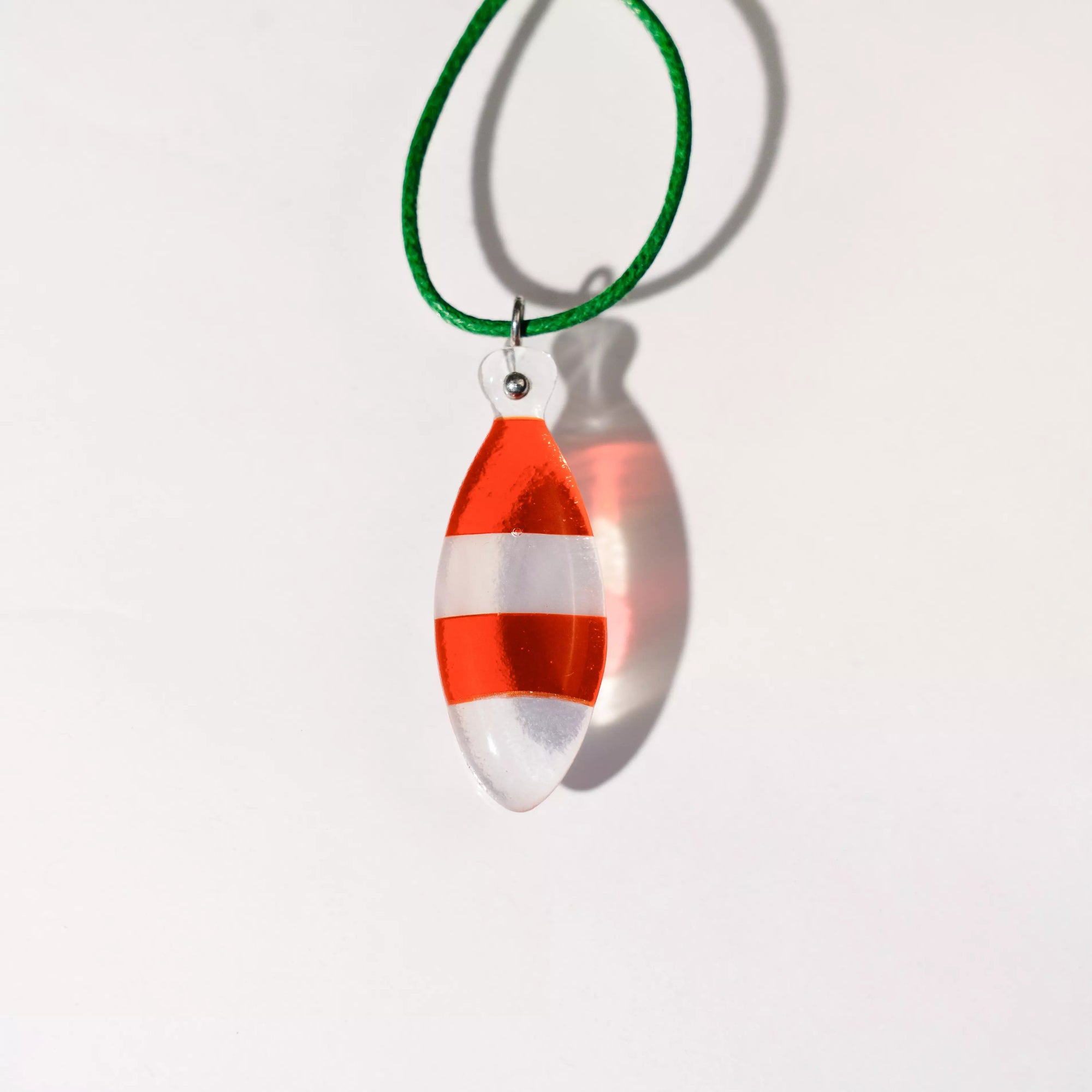 Coral-colored fused glass fish pendant with bold red and white stripes, casting a colorful shadow against a white backdrop.