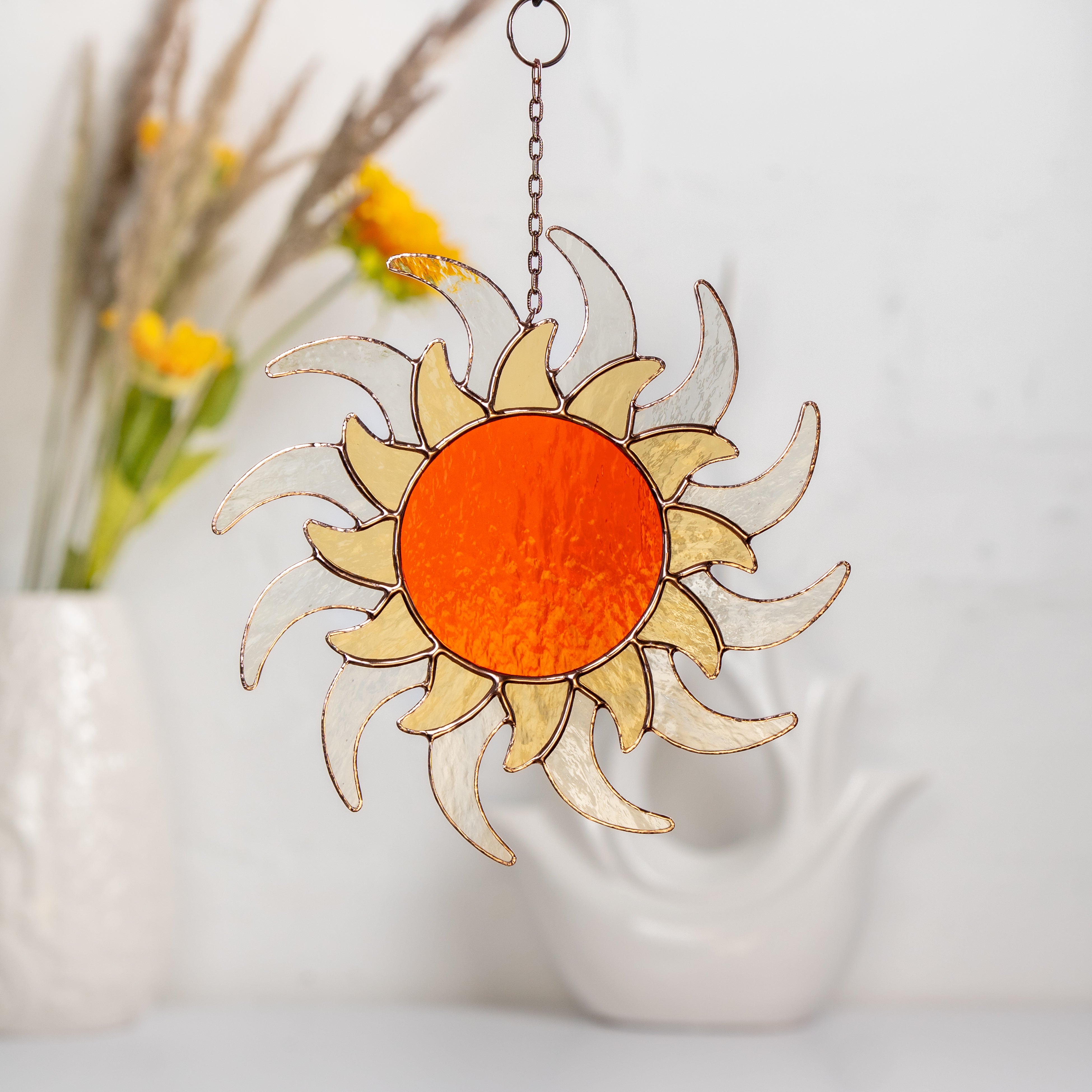 Stained glass sun hanging in interior with orange center and translucent rays, decorative vase in background.