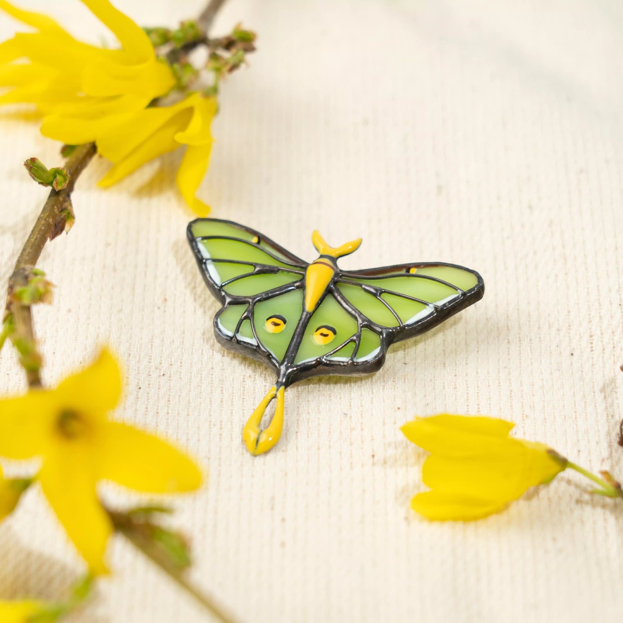 Butterfly stained glass green and yellow enamel pin on a textured surface with yellow flowers.