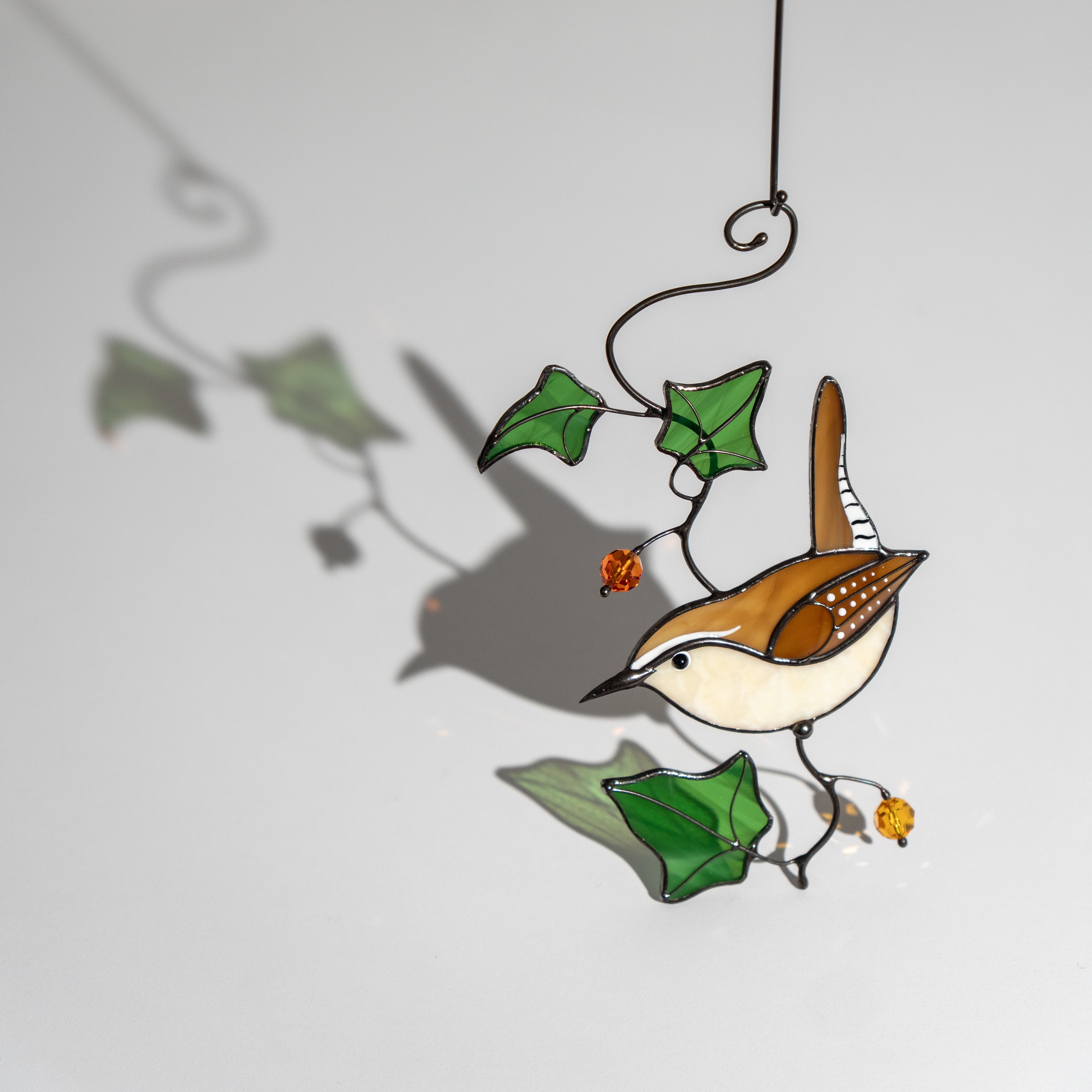 Carolina wren stained glass suncatcher with green ivy leaves and orange beads, casting a soft shadow on a light wall.
