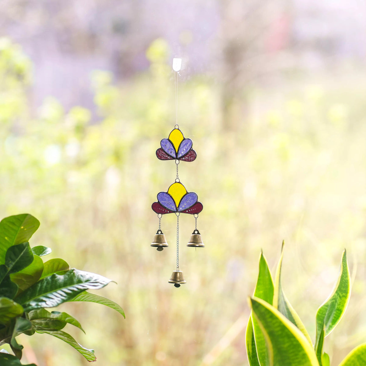 Close-up view of a colorful stained glass lotus wind chime near a window with lush green plants in the background.
