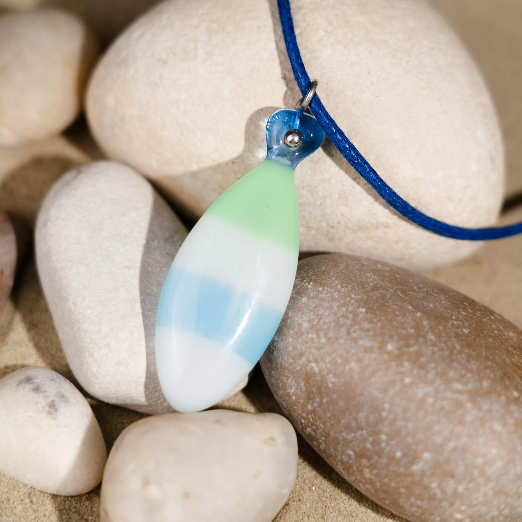 Fused glass fish pendant in Sea Breeze color palette resting on natural stones, with visible texture and soft light.