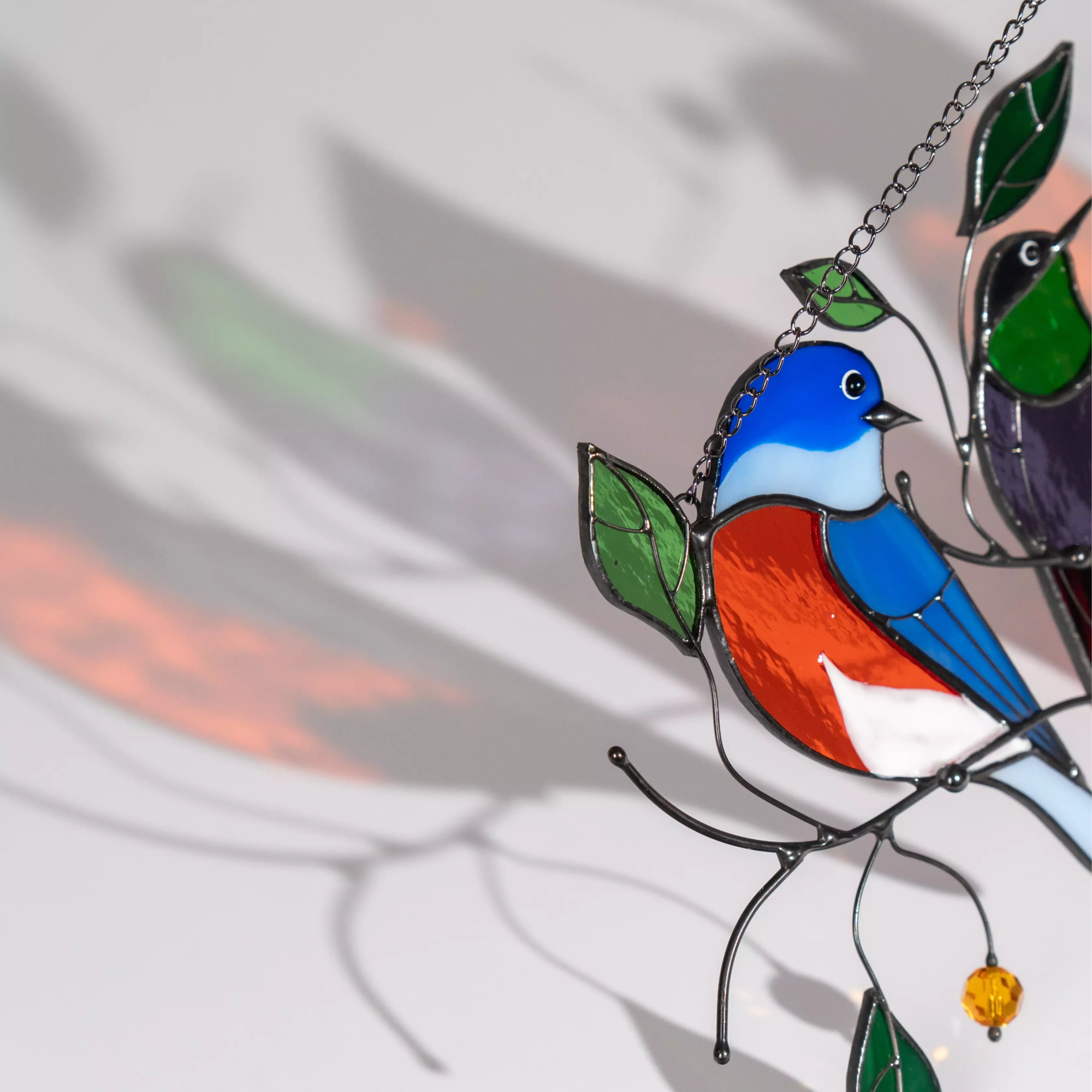 Close-up of stained glass bluebird on a branch with green leaves, casting a soft shadow against a light background