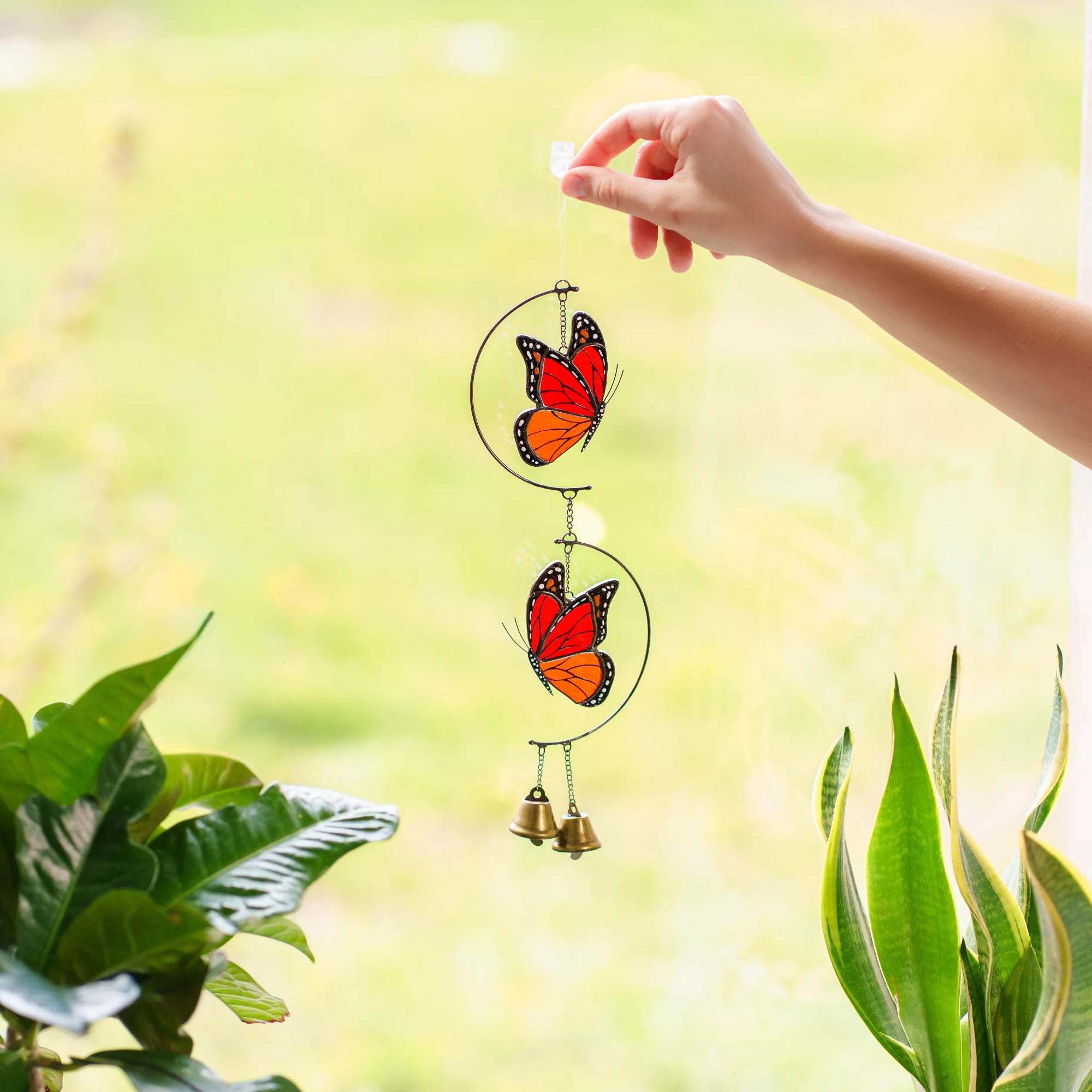 Close-up view of a person holding a stained glass wind chime with two monarch butterflies and brass bells near a window with green house plants.