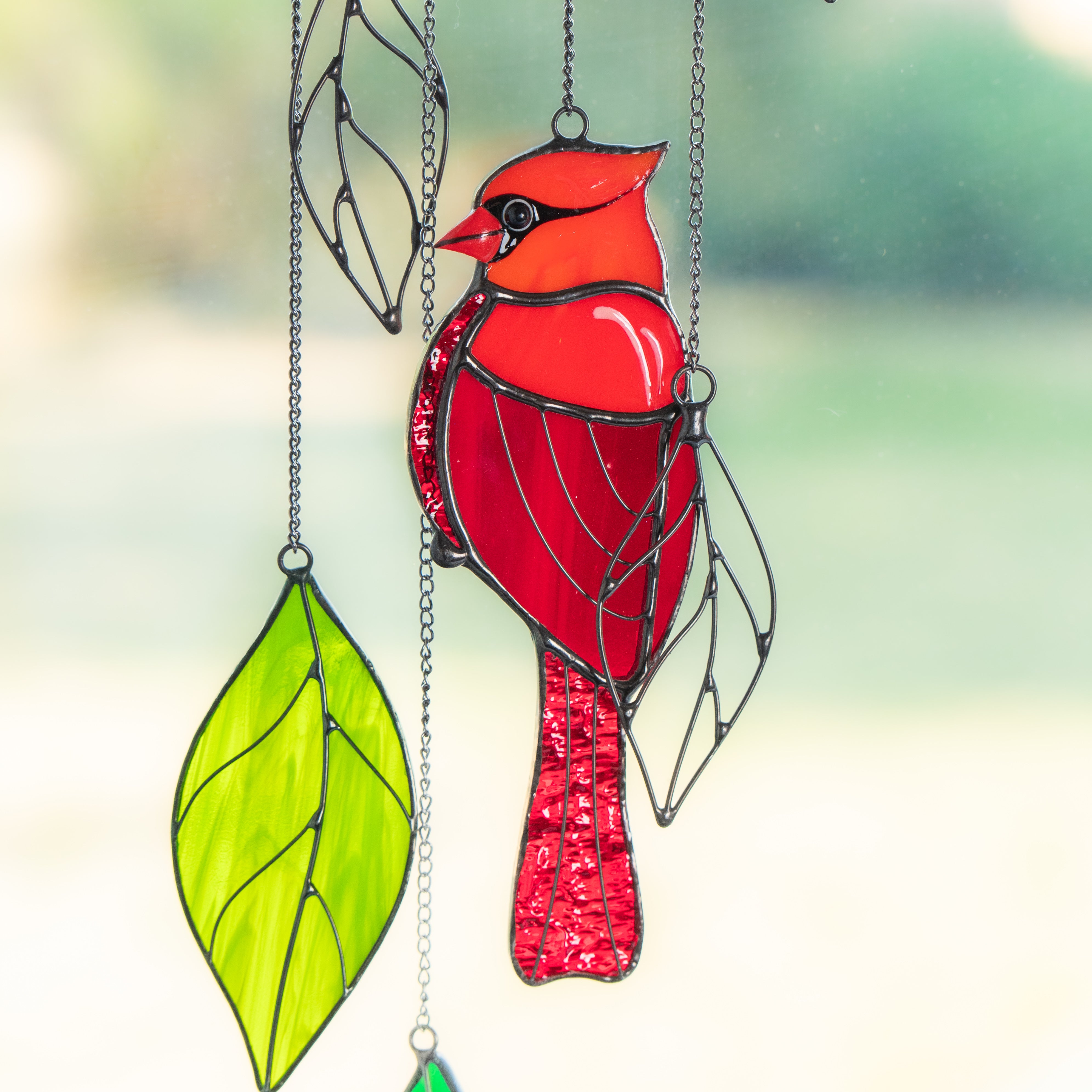 Bright red stained glass cardinal with vivid leaf ornaments hanging from a chain in the sunlight.
