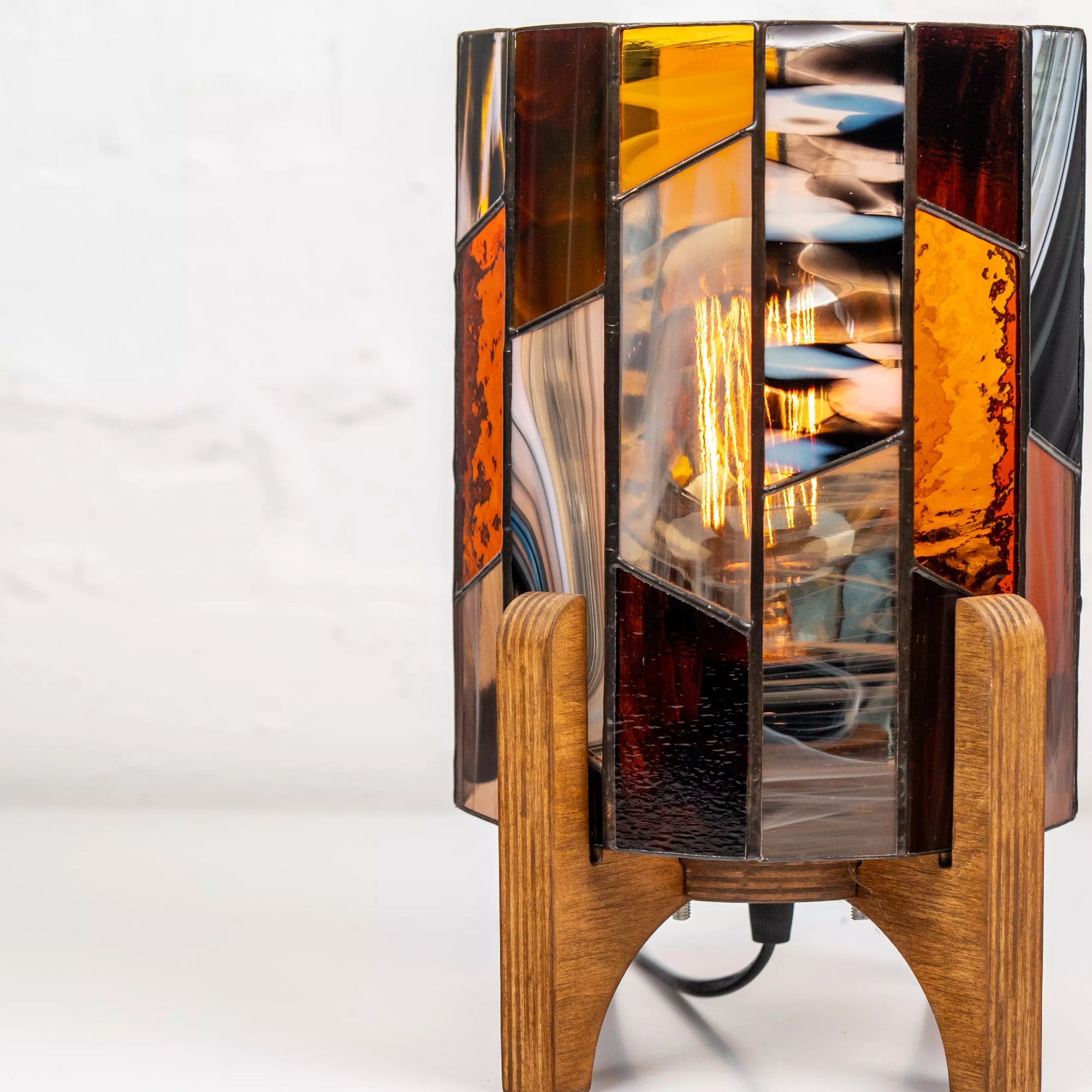 Close-up of handmade stained glass lamp with warm orange, gray, and amber glass textures on a solid wooden base, glowing softly.
