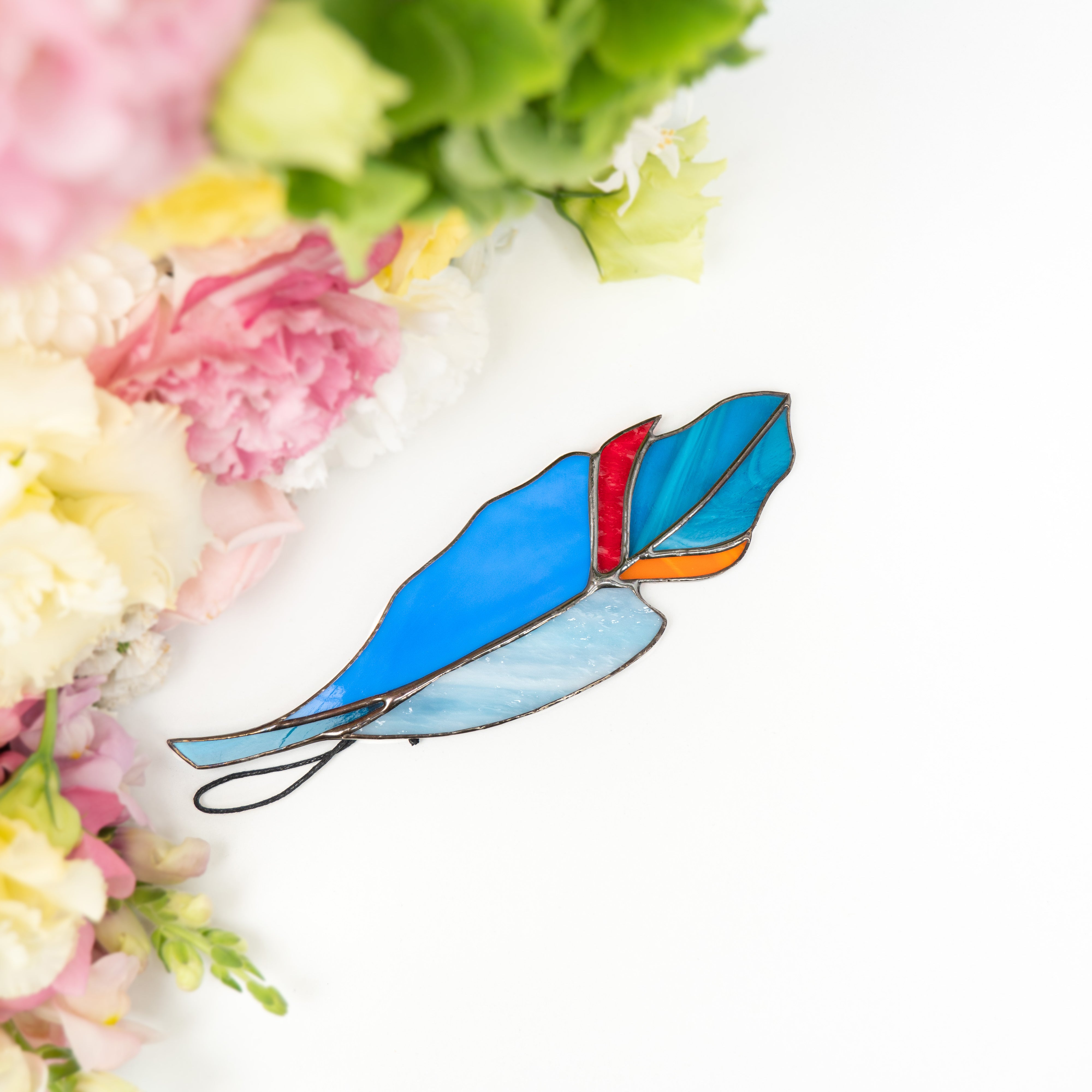 Feather suncatcher in shades of blue, red, and orange resting flat with soft pink and white flower petals.