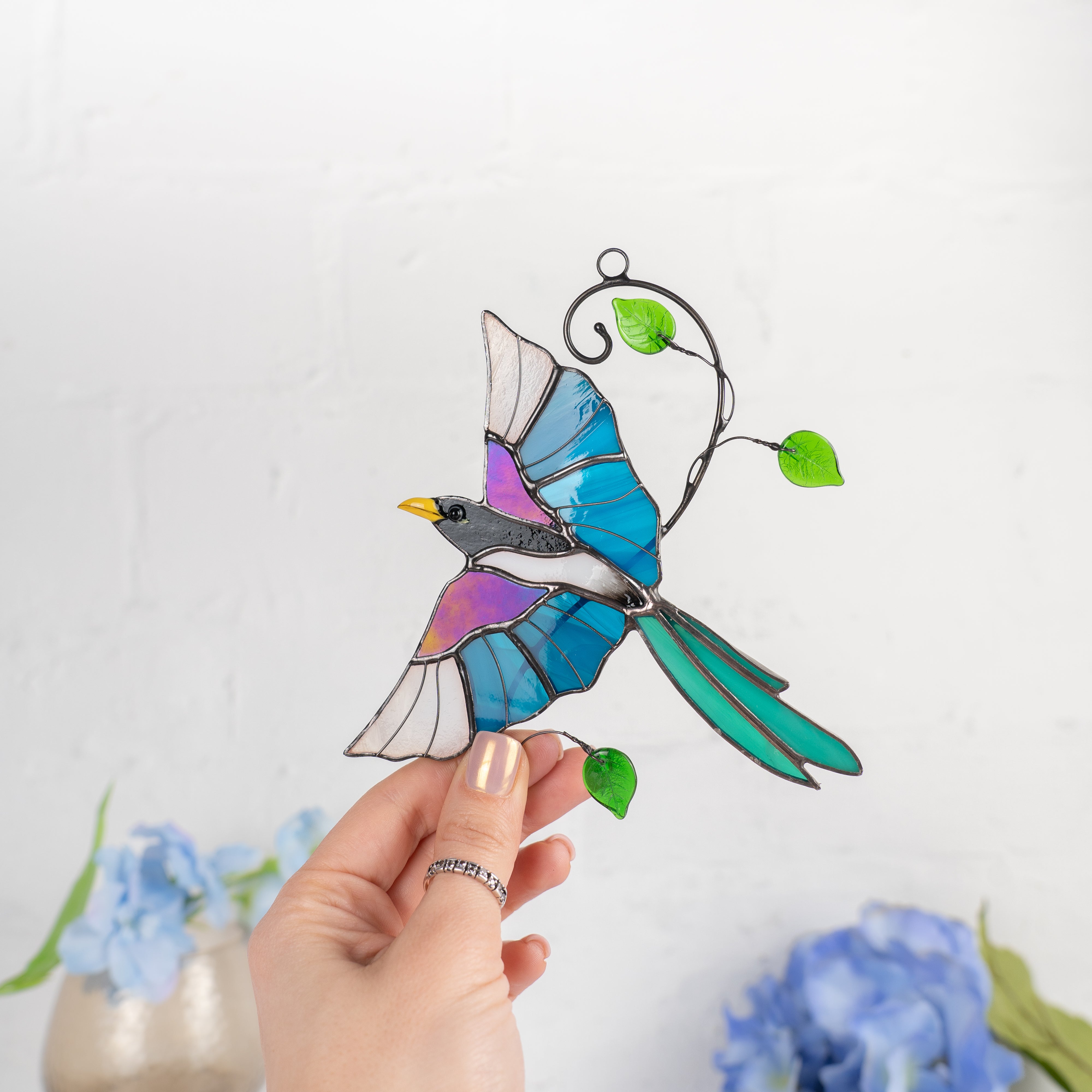 Hand holding a stained glass magpie with blue, purple, and white wings and a yellow beak in a bright interior.