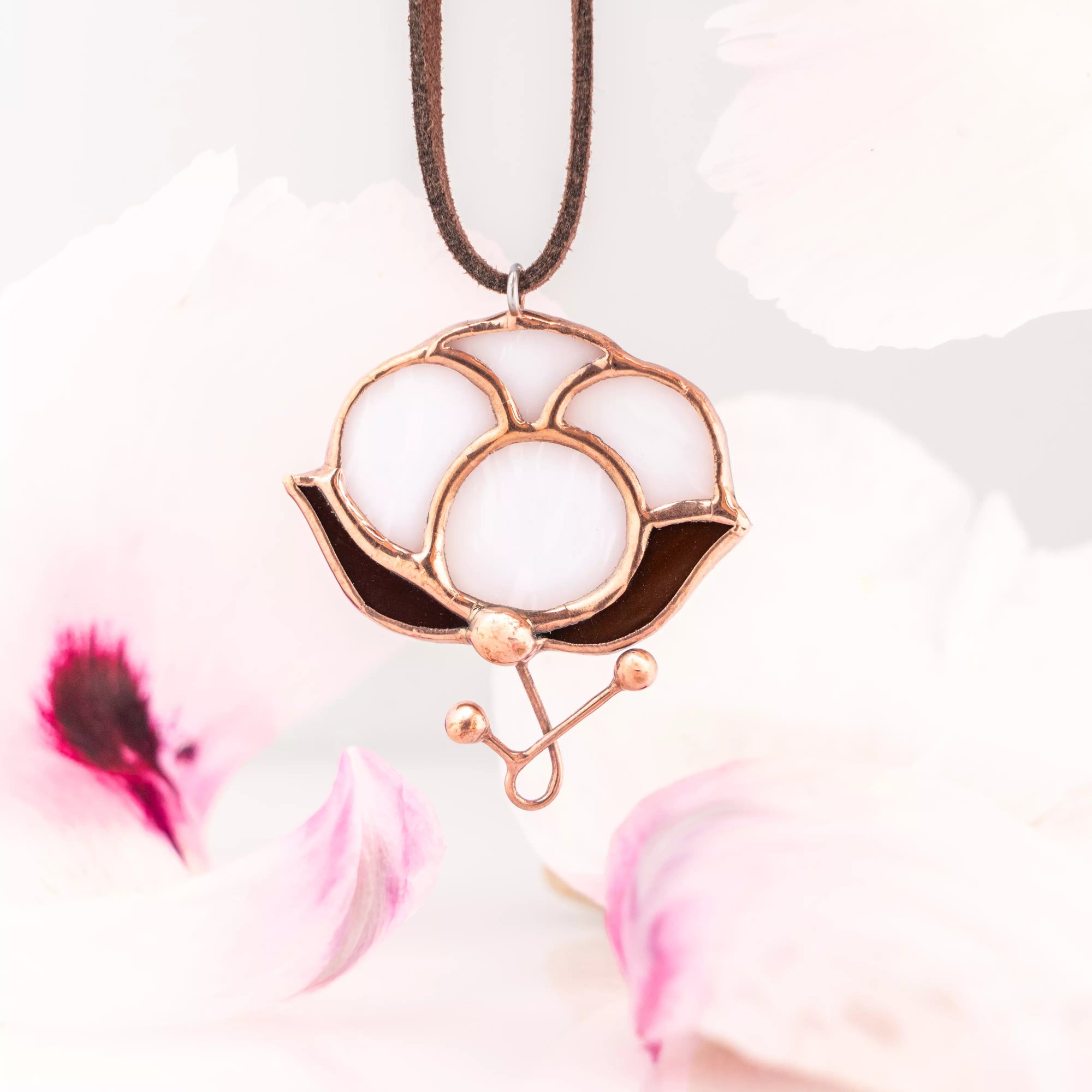Close-up of a white stained glass pendant shaped like a cotton flower with copper outlines, hanging on a suede cord among soft white petals.