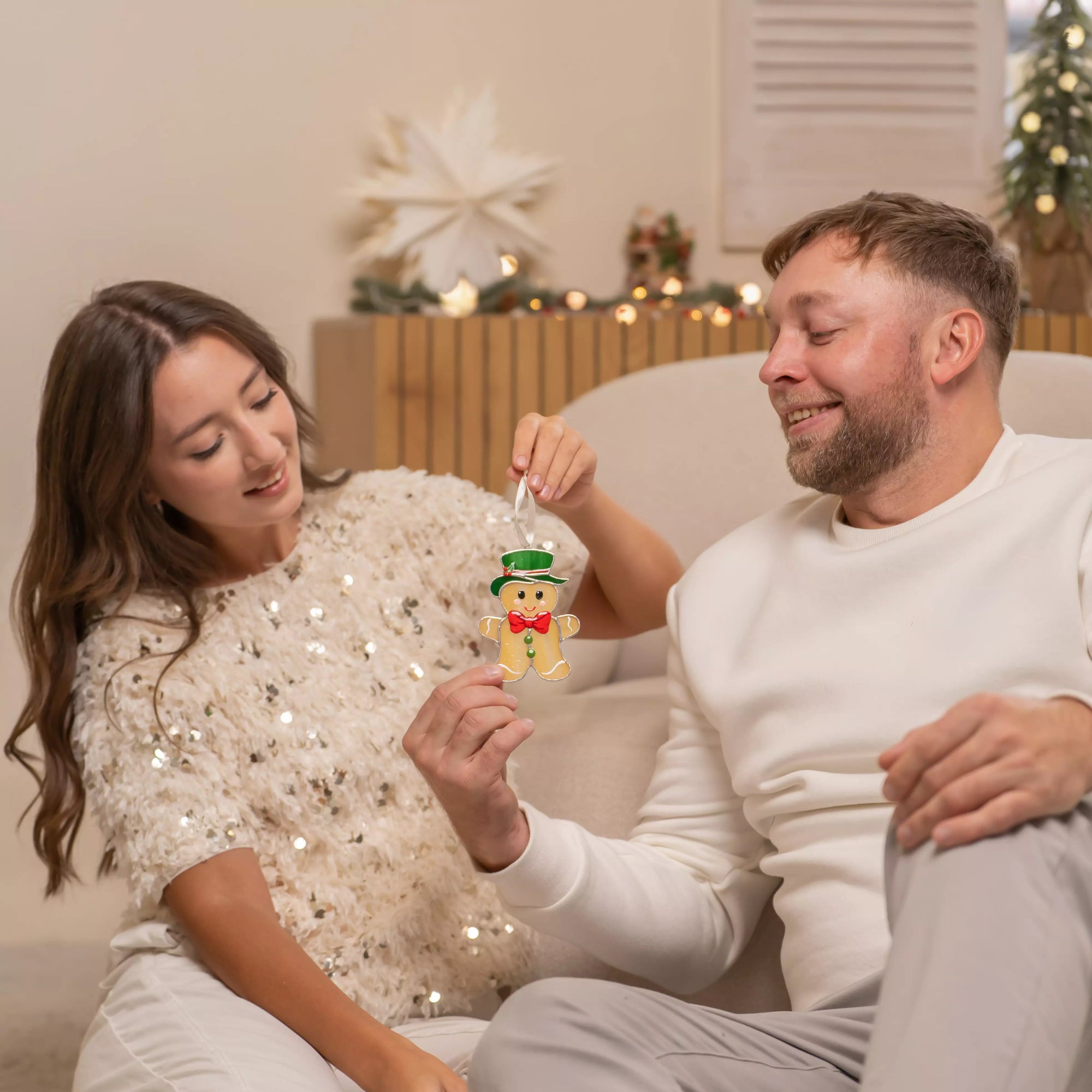 Smiling couple sitting together holding a stained glass gingerbread man ornament in a cozy holiday room.