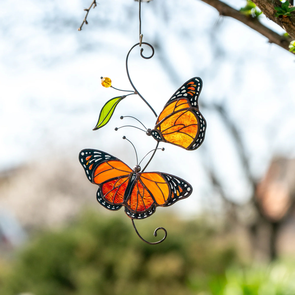 Monarch butterfly stained glass suncatcher hanging outdoors from a bare tree branch against a pale winter sky - handmade orange and amber wings with a green glass leaf