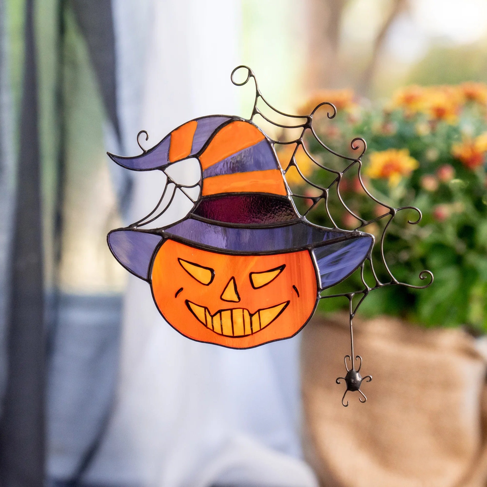 Stained glass pumpkin-in-a-witch-hat suncatcher glowing in a sunlit window with sheer curtains and orange autumn mums - handmade grinning jack-o'-lantern Halloween window decor with a spider web