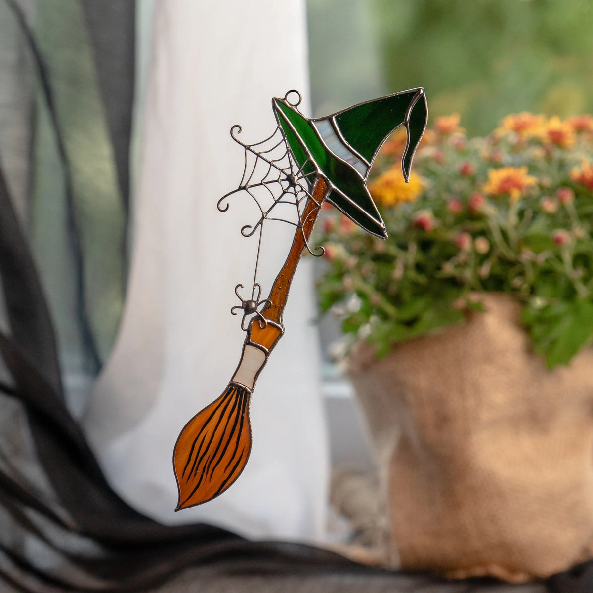 Witch hat and broom stained glass suncatcher in a window with black tulle and orange chrysanthemums - handmade Halloween decor for a windowsill through the whole season