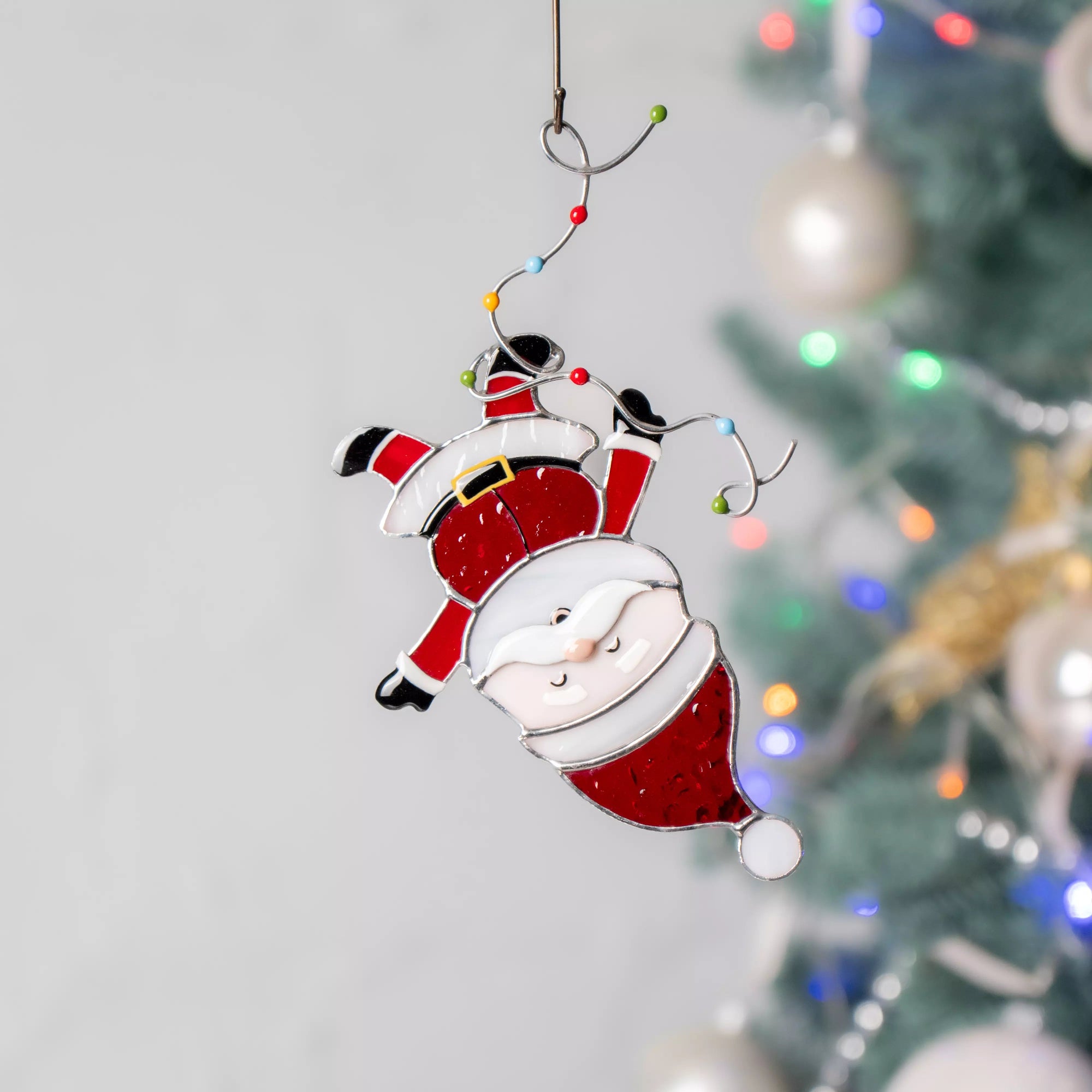 Stained glass Santa Claus suncatcher hanging against a blurred Christmas tree background.