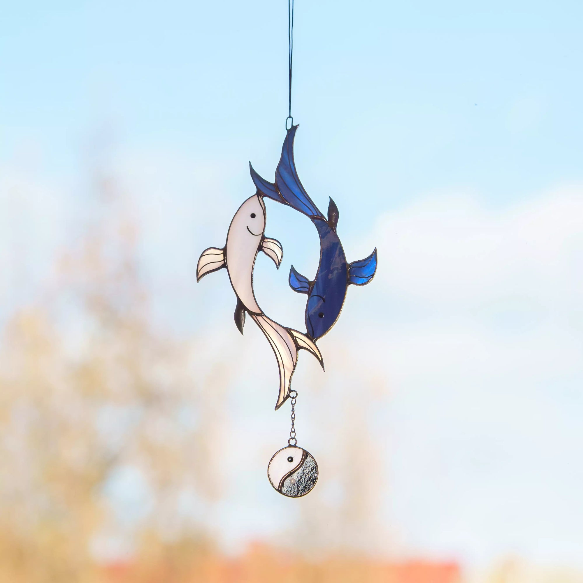 A blue and white koi fish stained glass suncatcher hanging against a soft, bright sky.