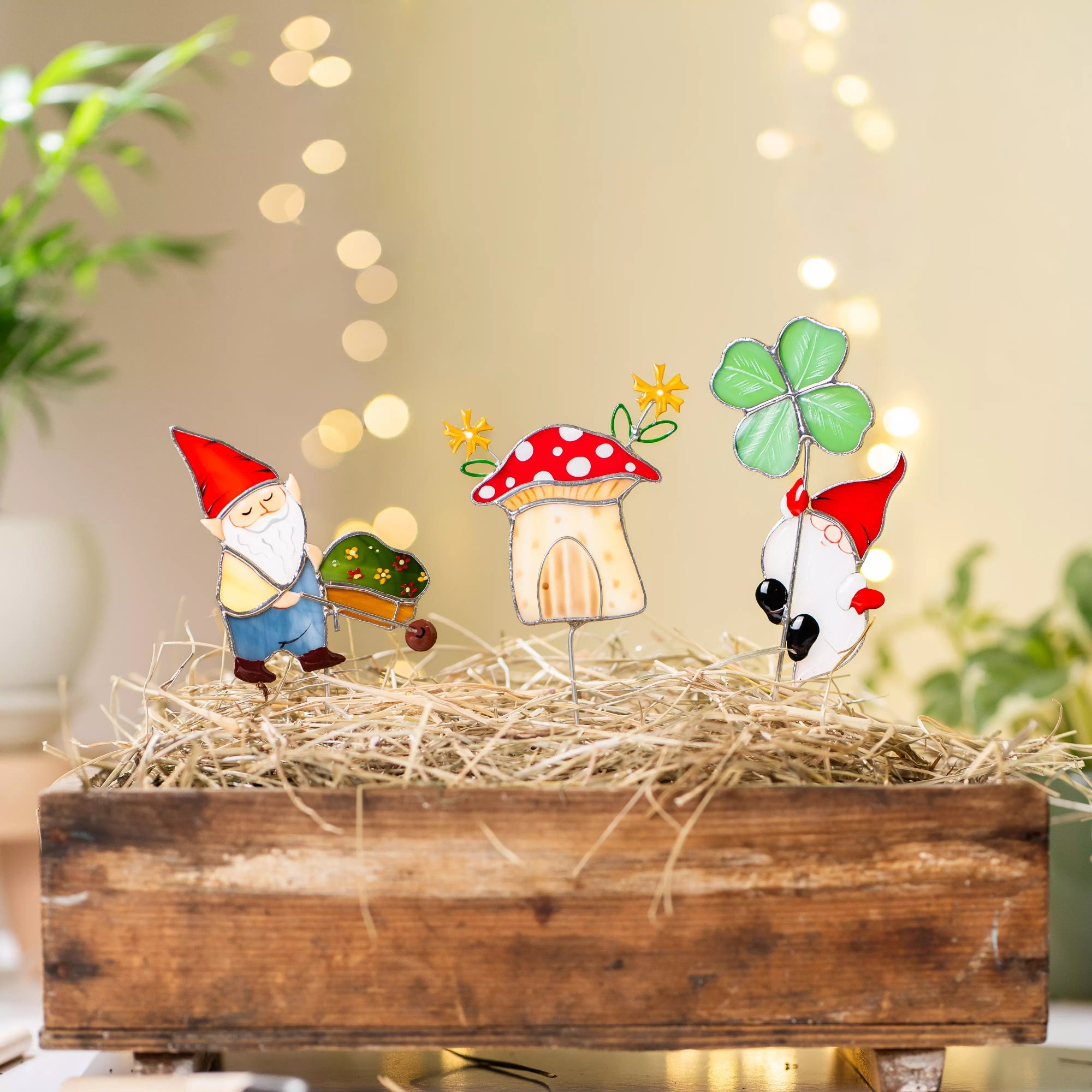 Stained glass garden gnome, mushroom, and bird plant stakes on a bed of straw with blurred lights in the background.