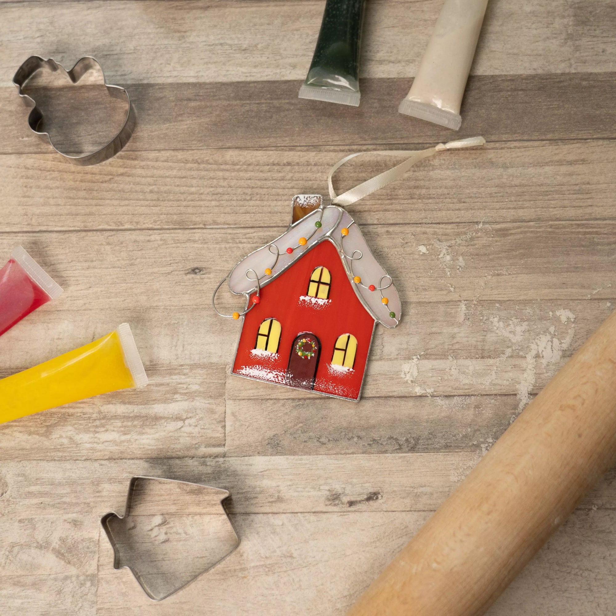 Stained glass gingerbread house ornament on a wooden surface with cookie cutters and a rolling pin.
