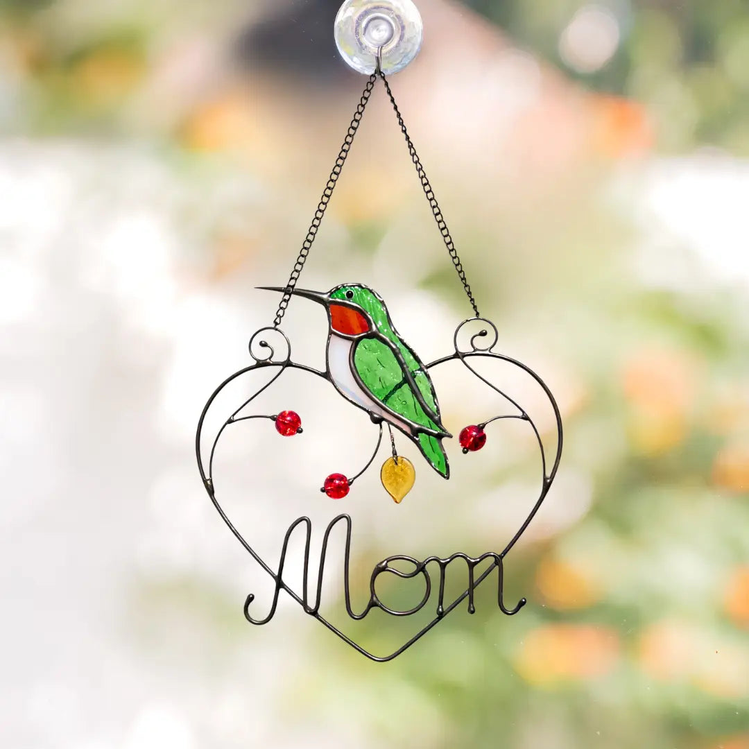 Decorative stained glass ornament with a green bird, red beads, and the word 'Mom' on a blurred natural background.