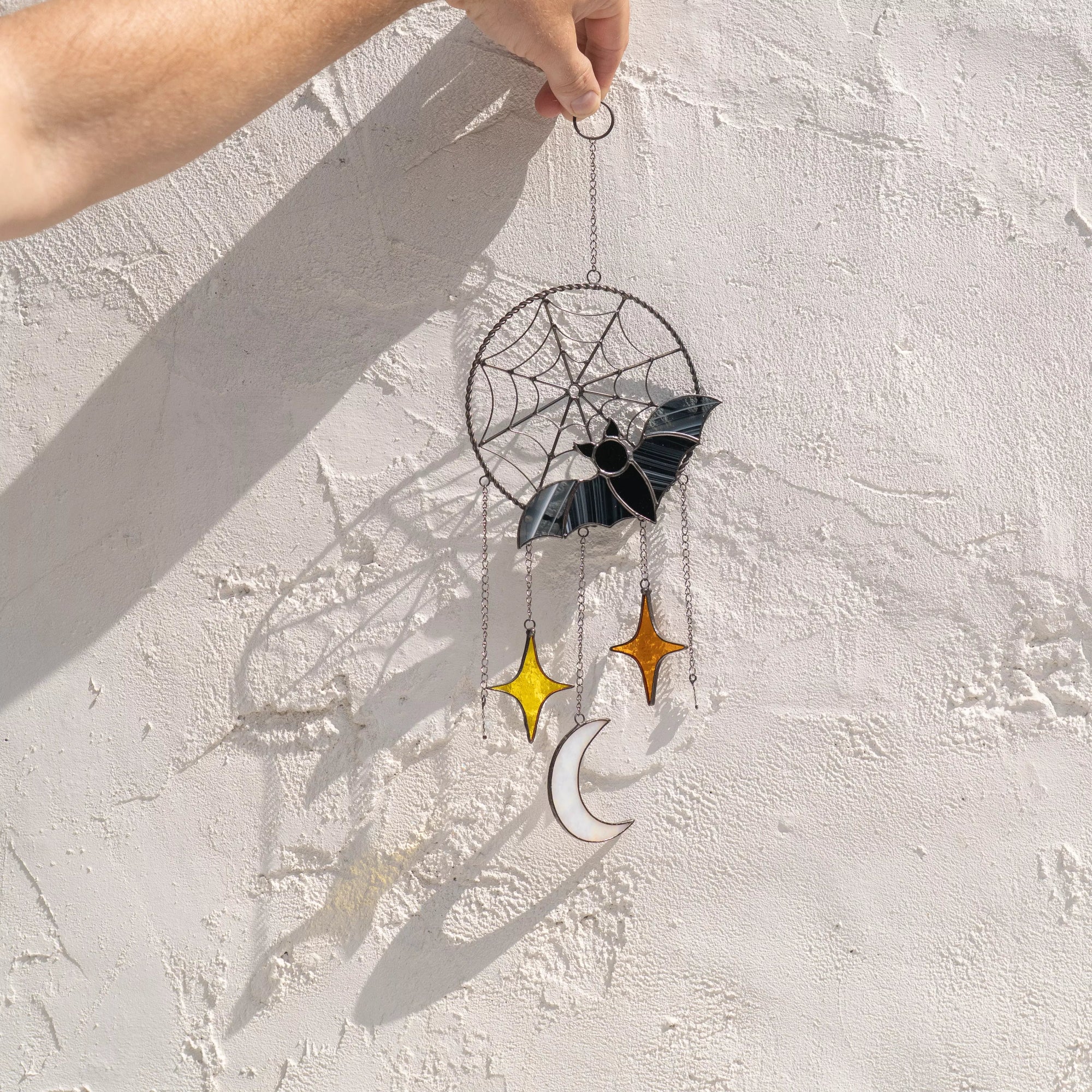 Decorative stained glass window hanging ornament with bat, star, and moon design casting a shadow on a textured white wall.