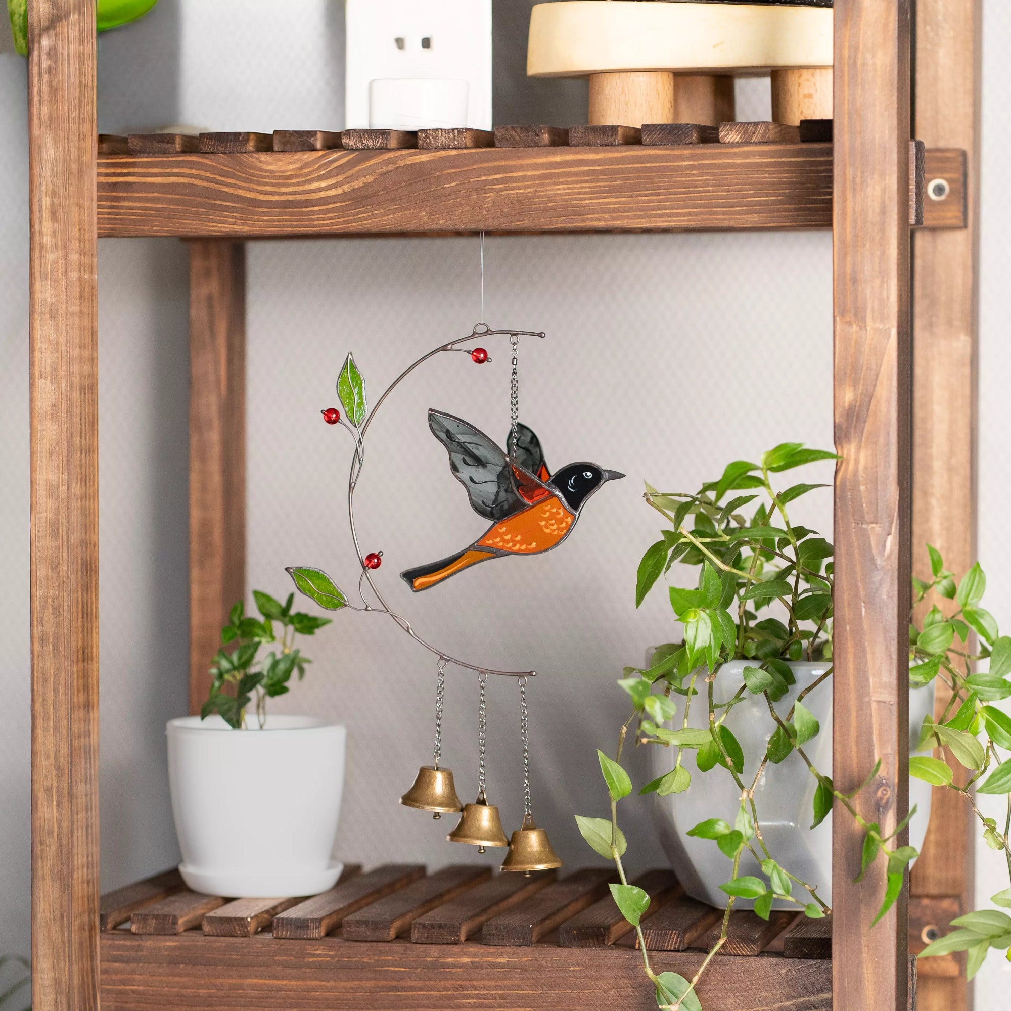 Decorative hanging ornament with baltimore oriole and bells on a wooden shelf with plants.