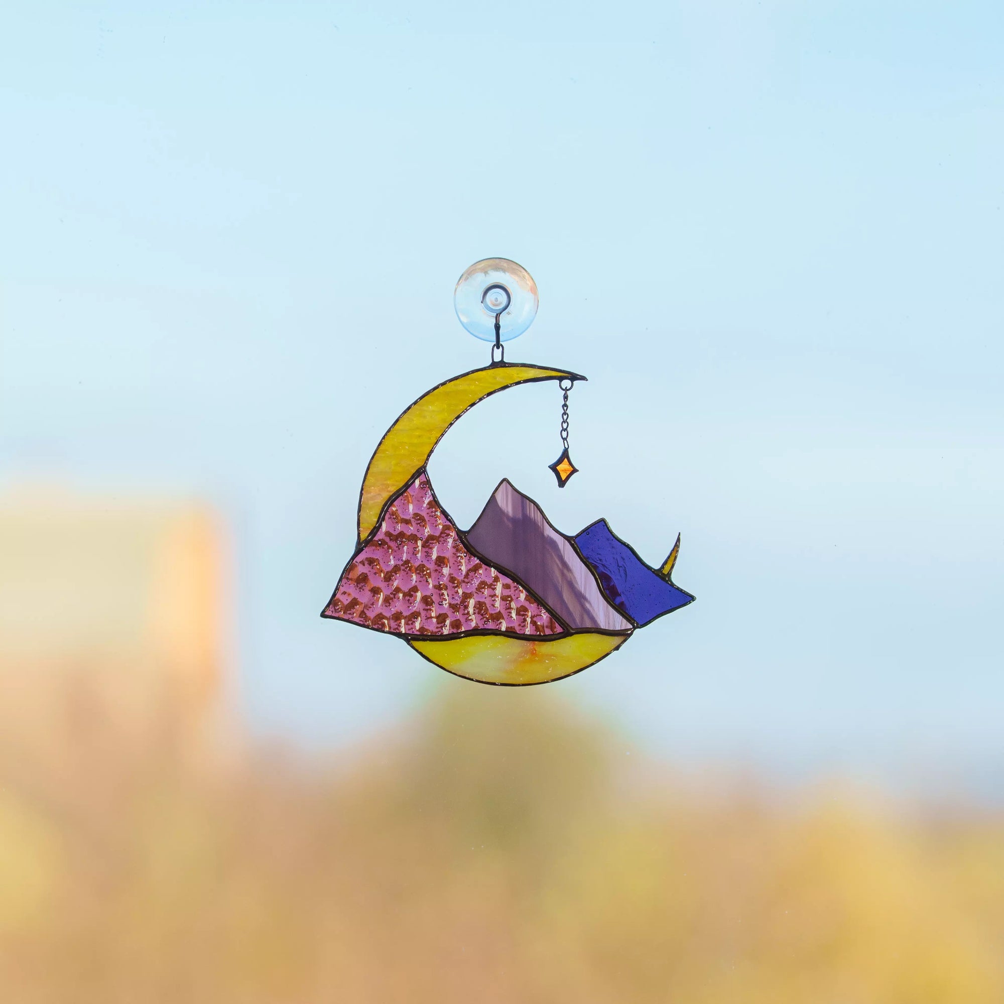 Stained glass window hanging with a colorful design of mountains and a crescent moon against a blurred natural background.