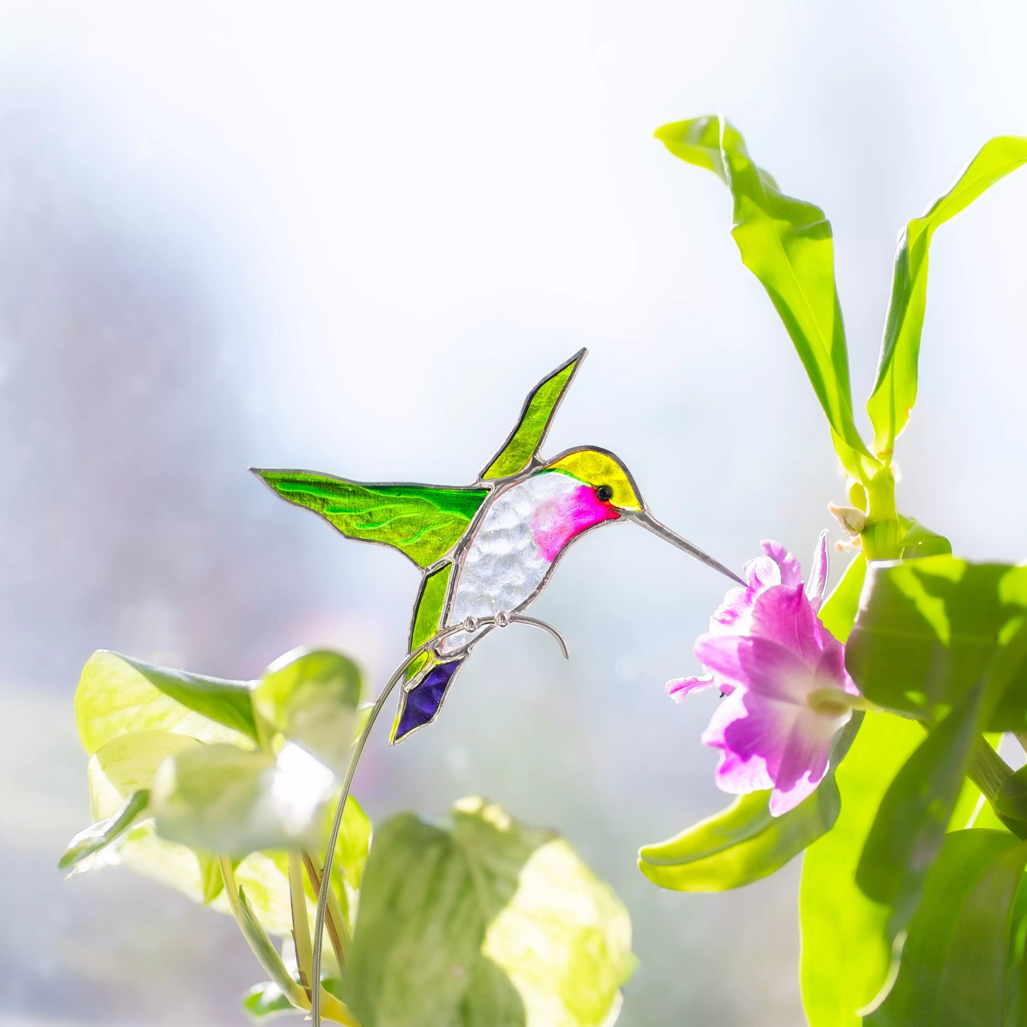 Stained glass hummingbird plant stake near a flower ornament on a branch with a blurred background
