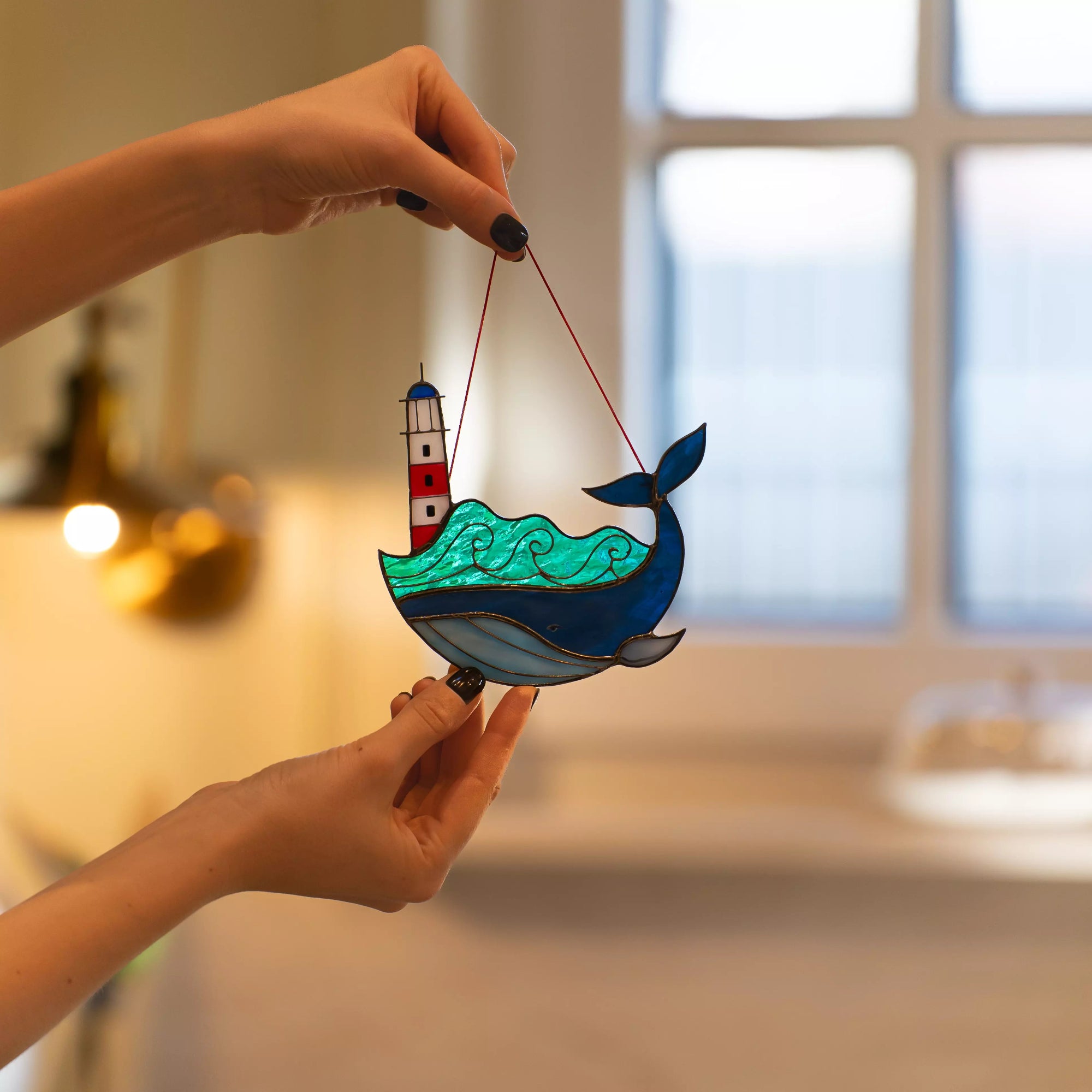 Stained glass suncatcher of a whale with a lighthouse held by a hand in a softly blurred indoor setting.