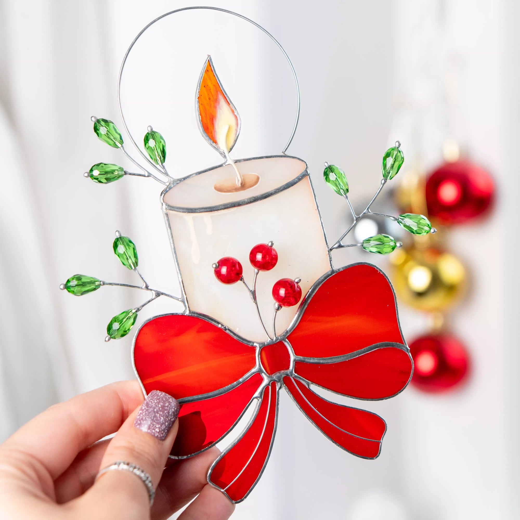 Stained glass ornament candle with red bow and beads held by a hand against a blurred festive background.