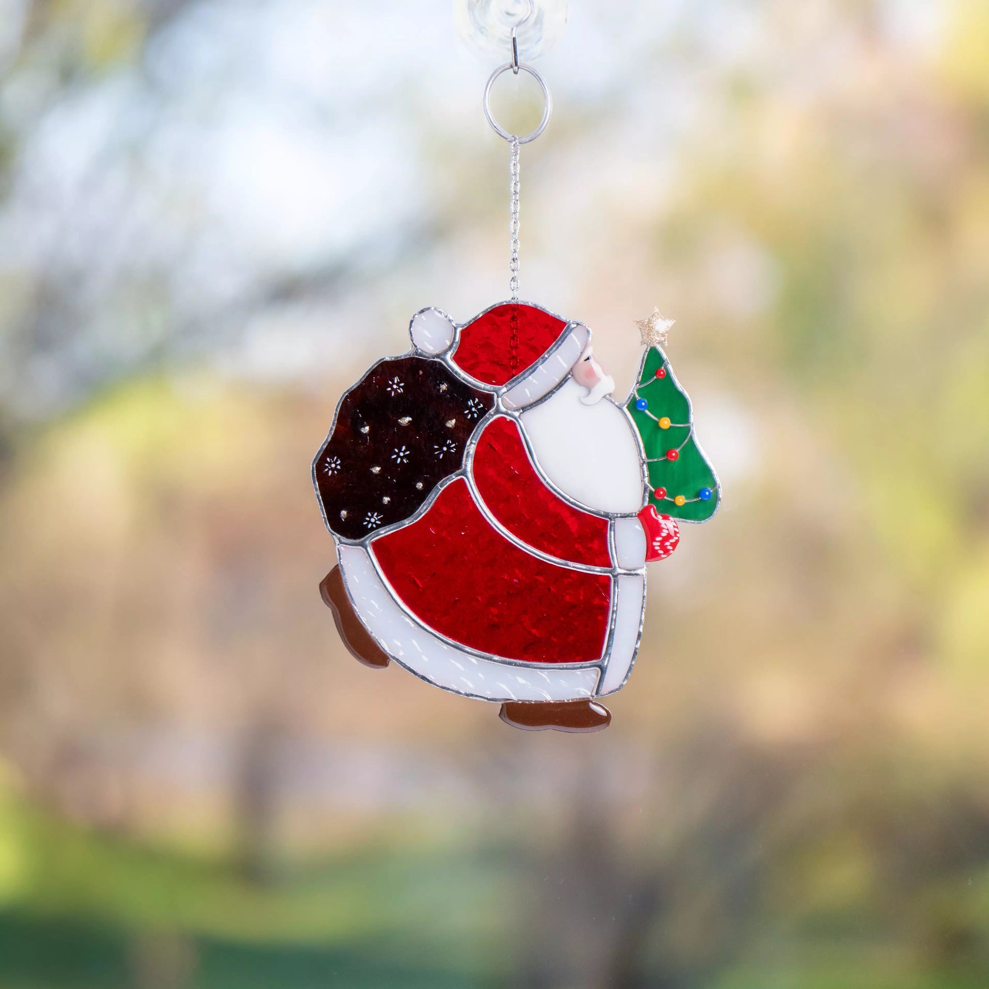 Stained glass suncatcher of a Santa Claus holding a Christmas tree against a blurred natural background.